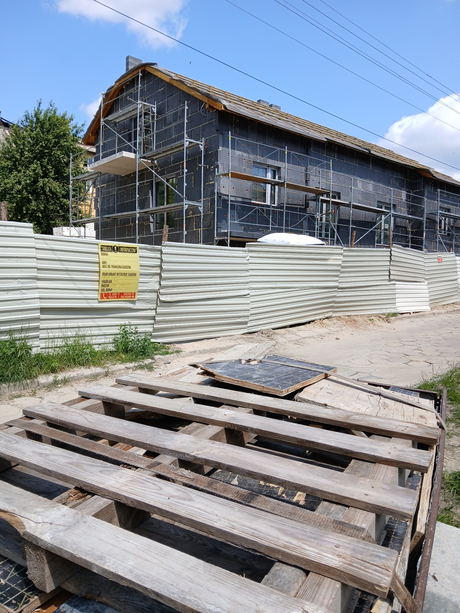 Dom w trakcie ocieplania, zabezpieczony rusztowaniami i ogrodzeniem. Widoczna tablica informacyjna budowy. Palety na pierwszym planie.