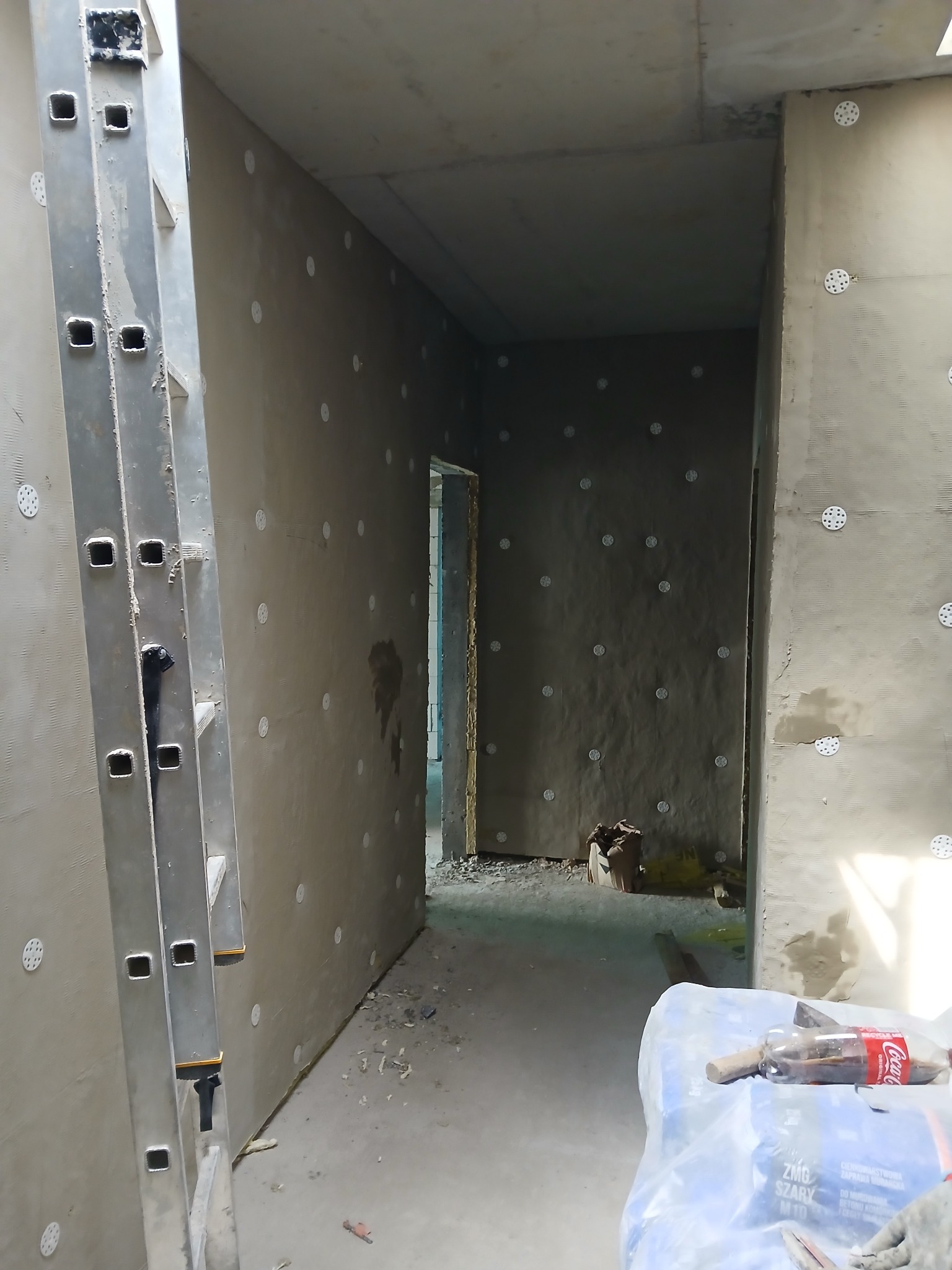 Wnętrze w trakcie ocieplania ścian styropianem, widoczne kołki mocujące, fragment drabiny aluminiowej i worki z zaprawą murarską w pomieszczeniu.