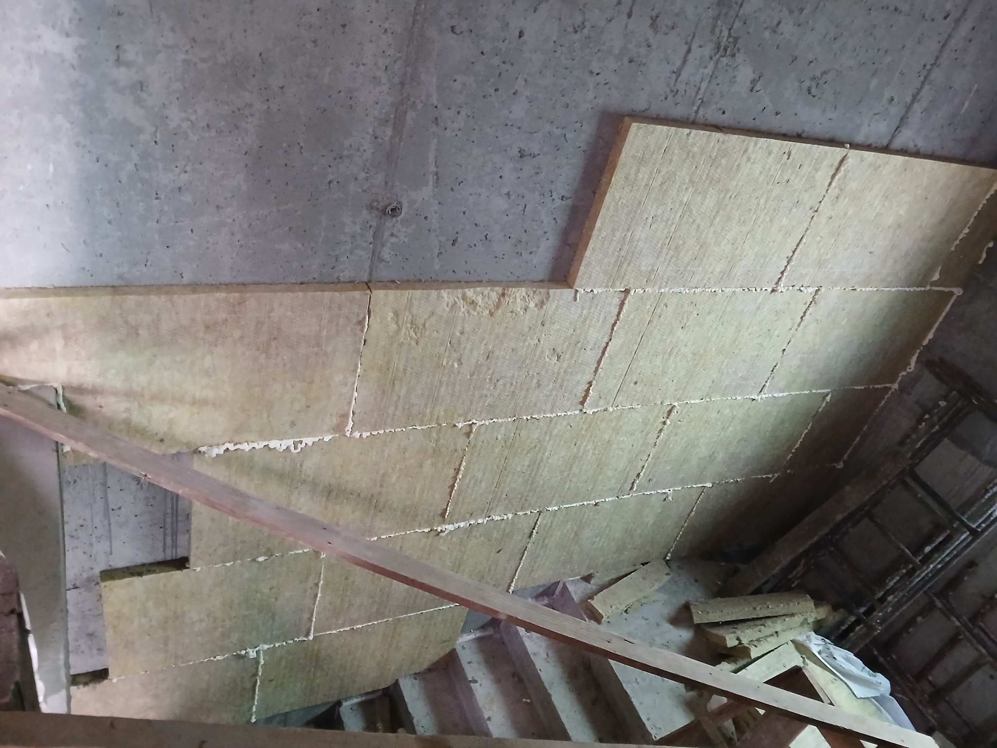 Ocieplenie wełną mineralną pod stropem, widoczne płyty izolacyjne z pianką montażową, fragment drewnianej konstrukcji i surowy beton. Prace izolacyjne w toku.