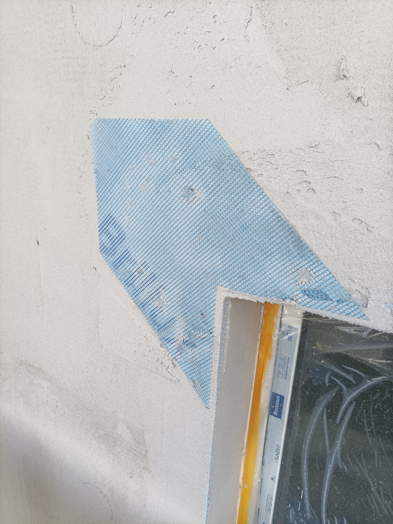 Fragment ściany z siatką elewacyjną przy oknie, przygotowanie do tynkowania. Widoczne zabezpieczenie okna folią i taśmą. Szorstka faktura ściany.