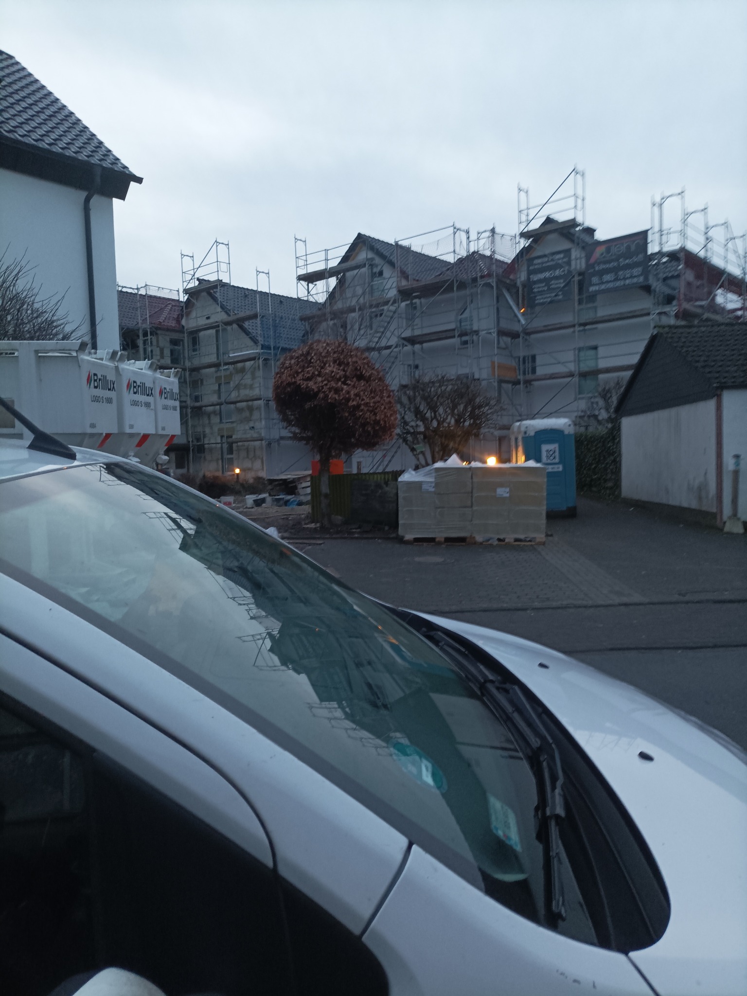 Domy w rusztowaniach z bliska, widoczne kontenery Brillux i przenośna toaleta. W tle zabudowa mieszkalna w trakcie prac elewacyjnych.