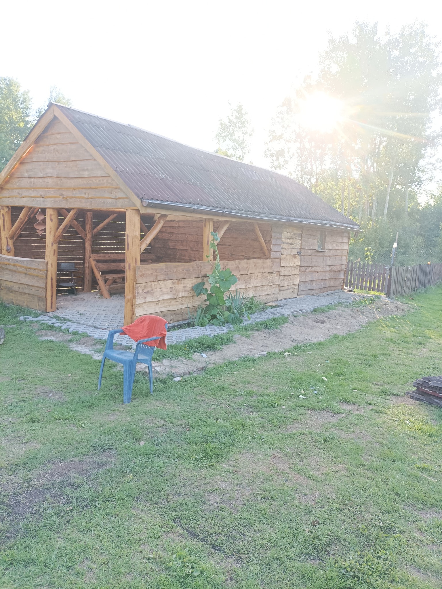 Drewniana altana z metalowym dachem, otoczona trawnikiem i ogrodzeniem. Na pierwszym planie plastikowe krzesło z czerwoną tkaniną. Słoneczny dzień, widoczne drzewa w tle.