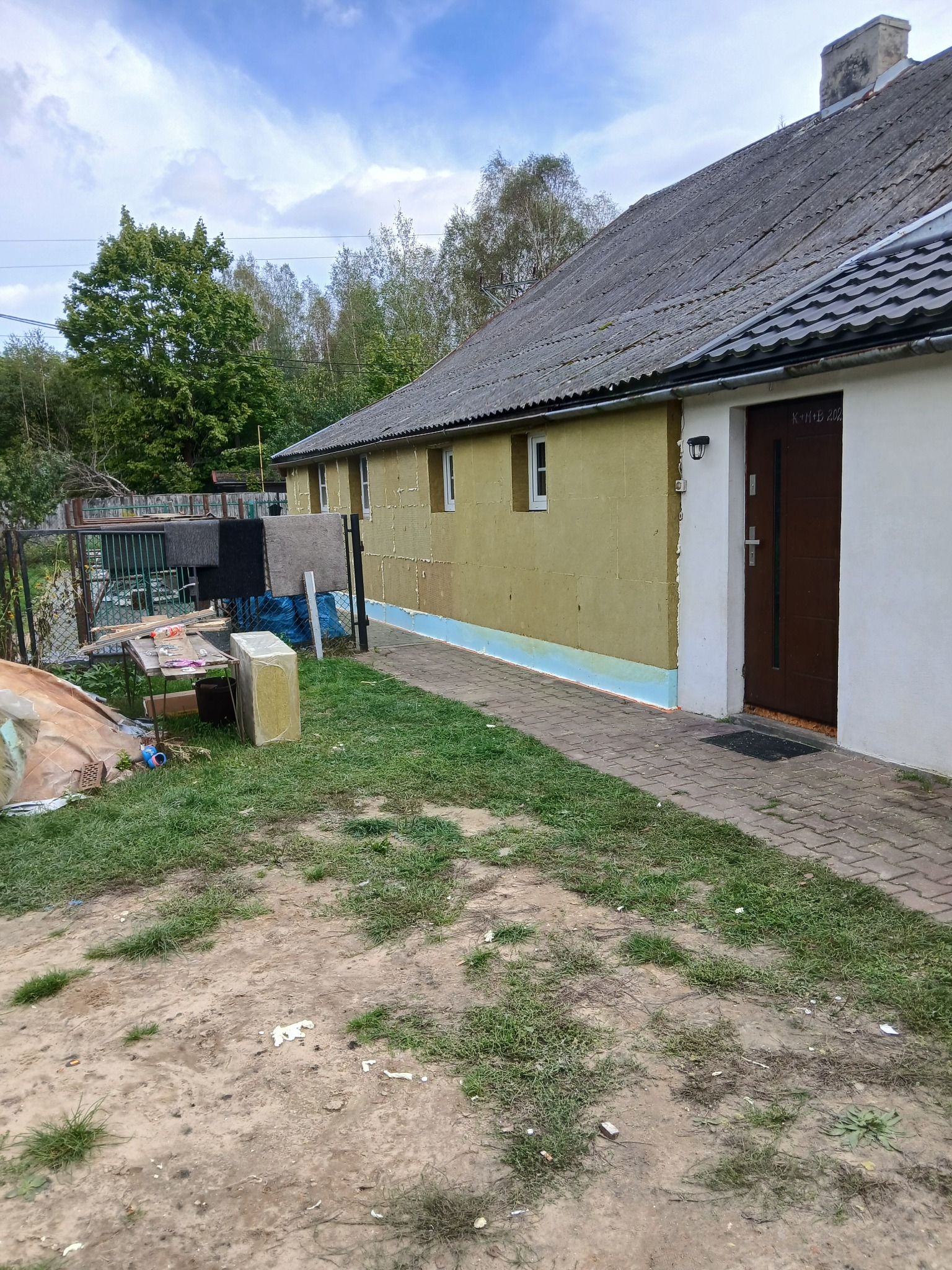 Dom w trakcie ocieplania: żółty styropian na elewacji, fragment otynkowanej ściany. Widoczne okna, drzwi wejściowe i elementy rusztowania. Prace w toku.