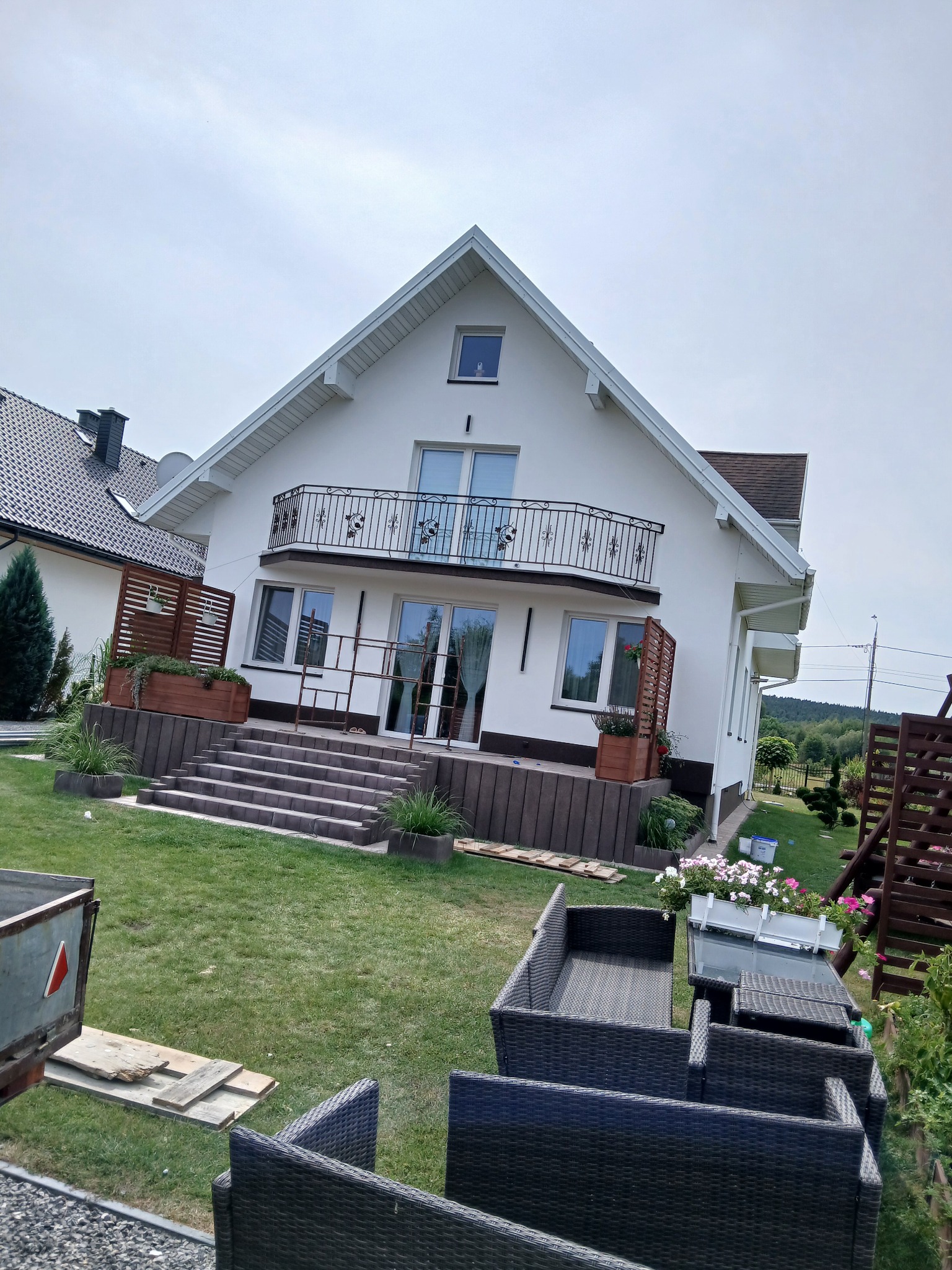 Biały dom z balkonem i schodami na tle zielonego trawnika. Widoczne meble ogrodowe i elementy dekoracyjne. Ujęcie z dołu podkreśla architekturę budynku.