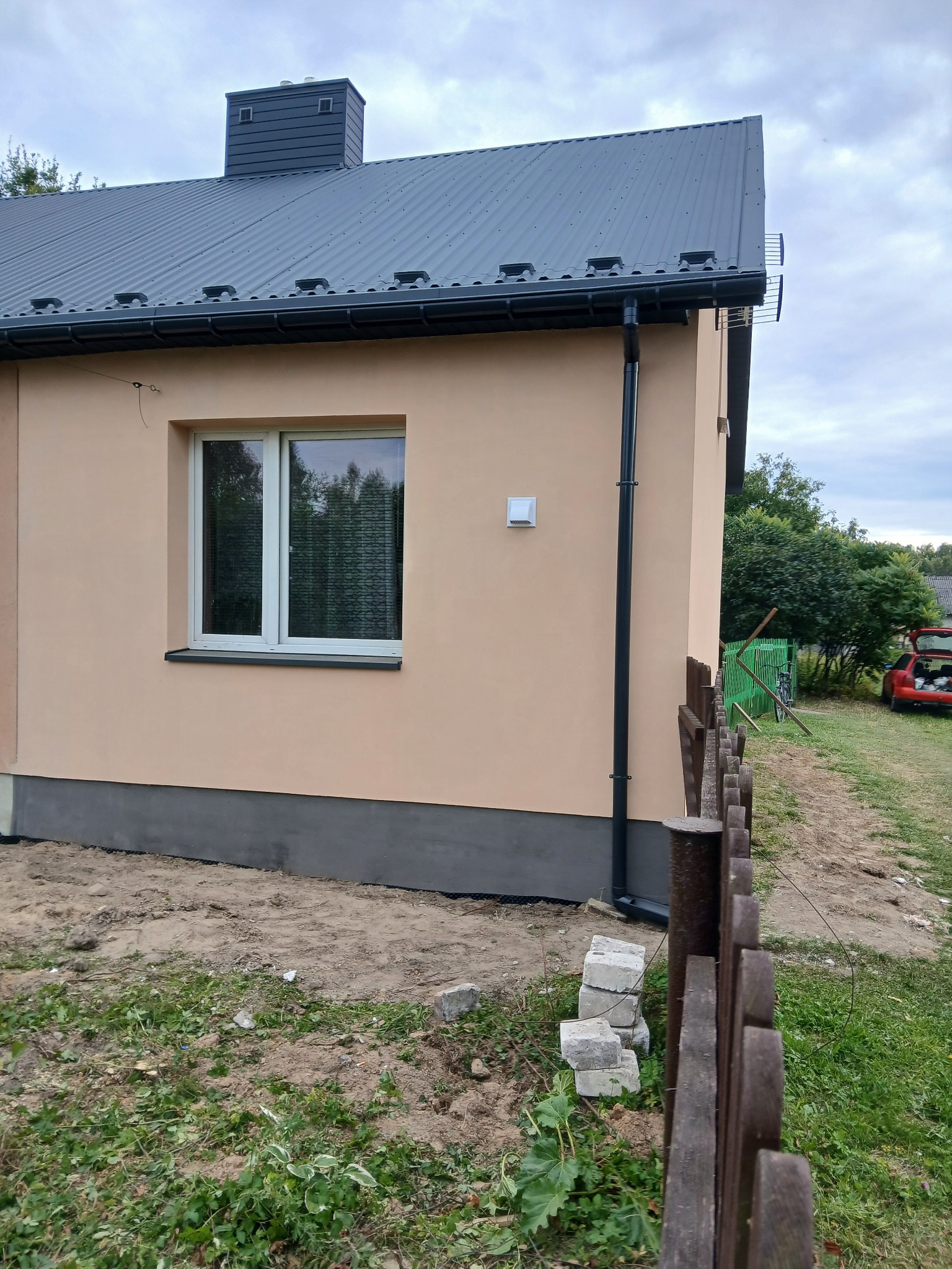 Wykończona elewacja domu jednorodzinnego z beżowym tynkiem, szarą podmurówką i grafitowym dachem. Widoczny fragment drewnianego płotu i ułożone bloczki.