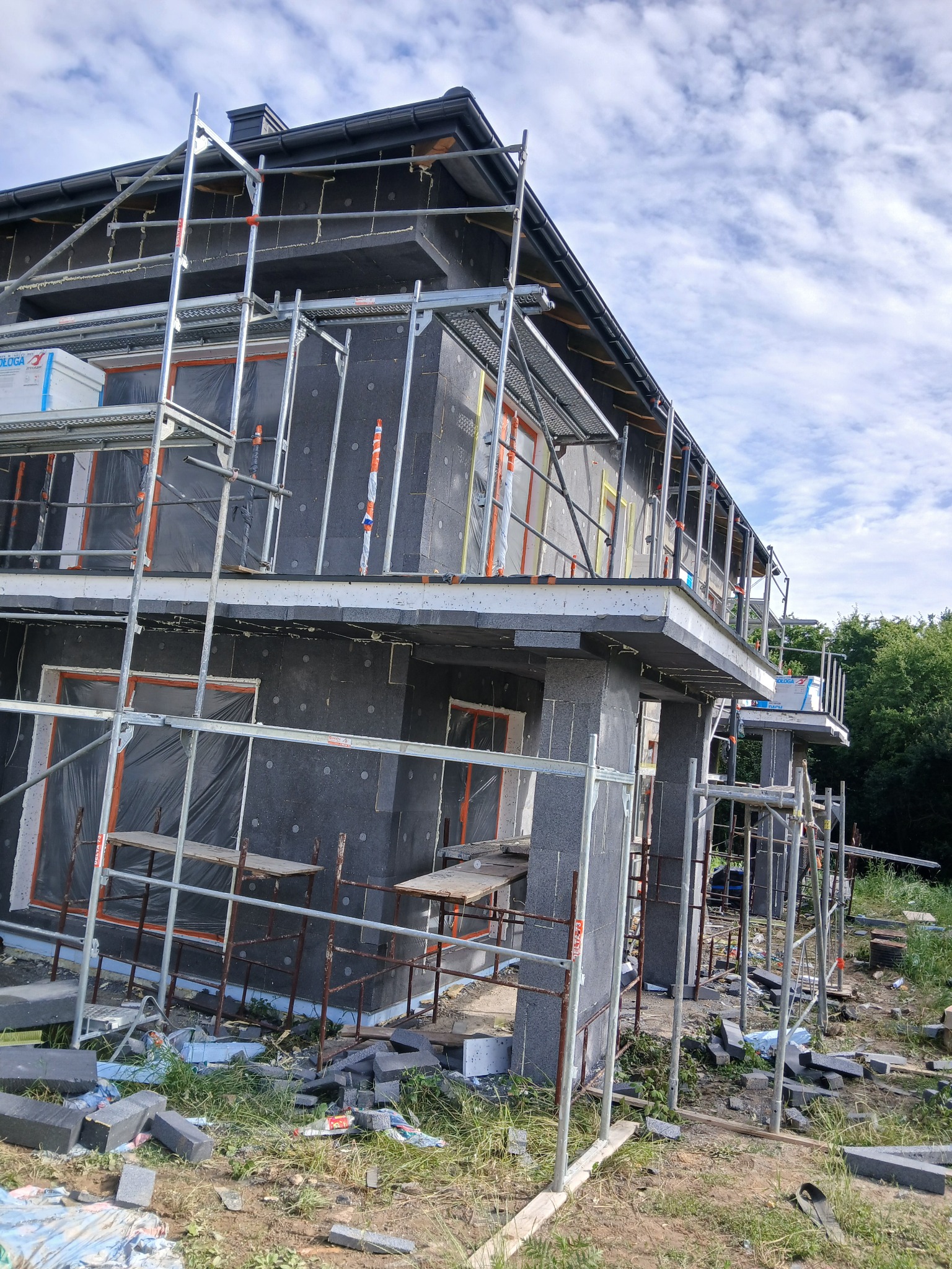 Dom w trakcie ocieplania elewacji styropianem, widoczne rusztowania i materiały budowlane. Prace postępują, okna zabezpieczone folią.