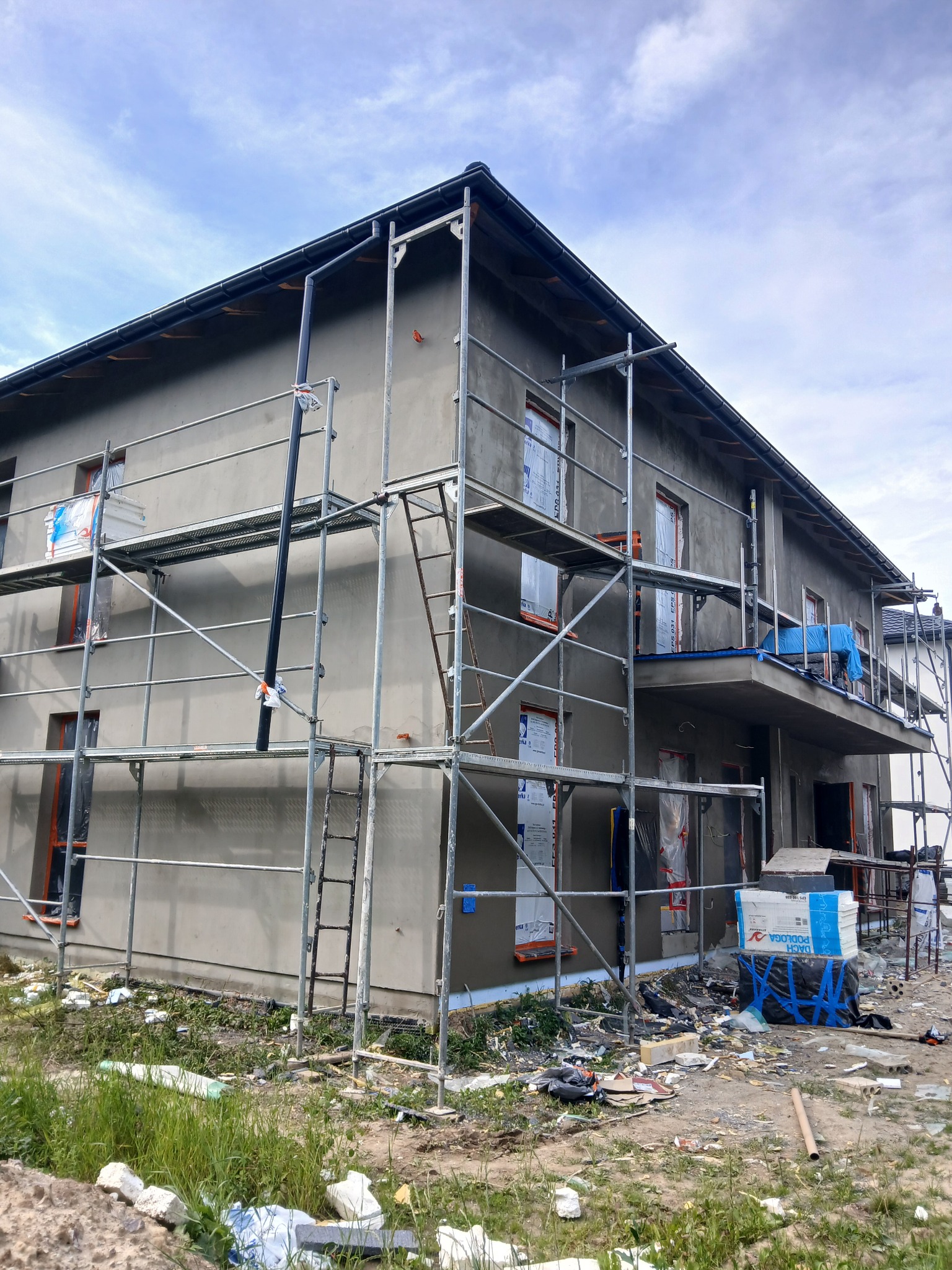 Dom w trakcie tynkowania elewacji, otoczony rusztowaniami. Okna zabezpieczone folią, widoczne materiały budowlane i elementy wykończeniowe. Prace w toku.