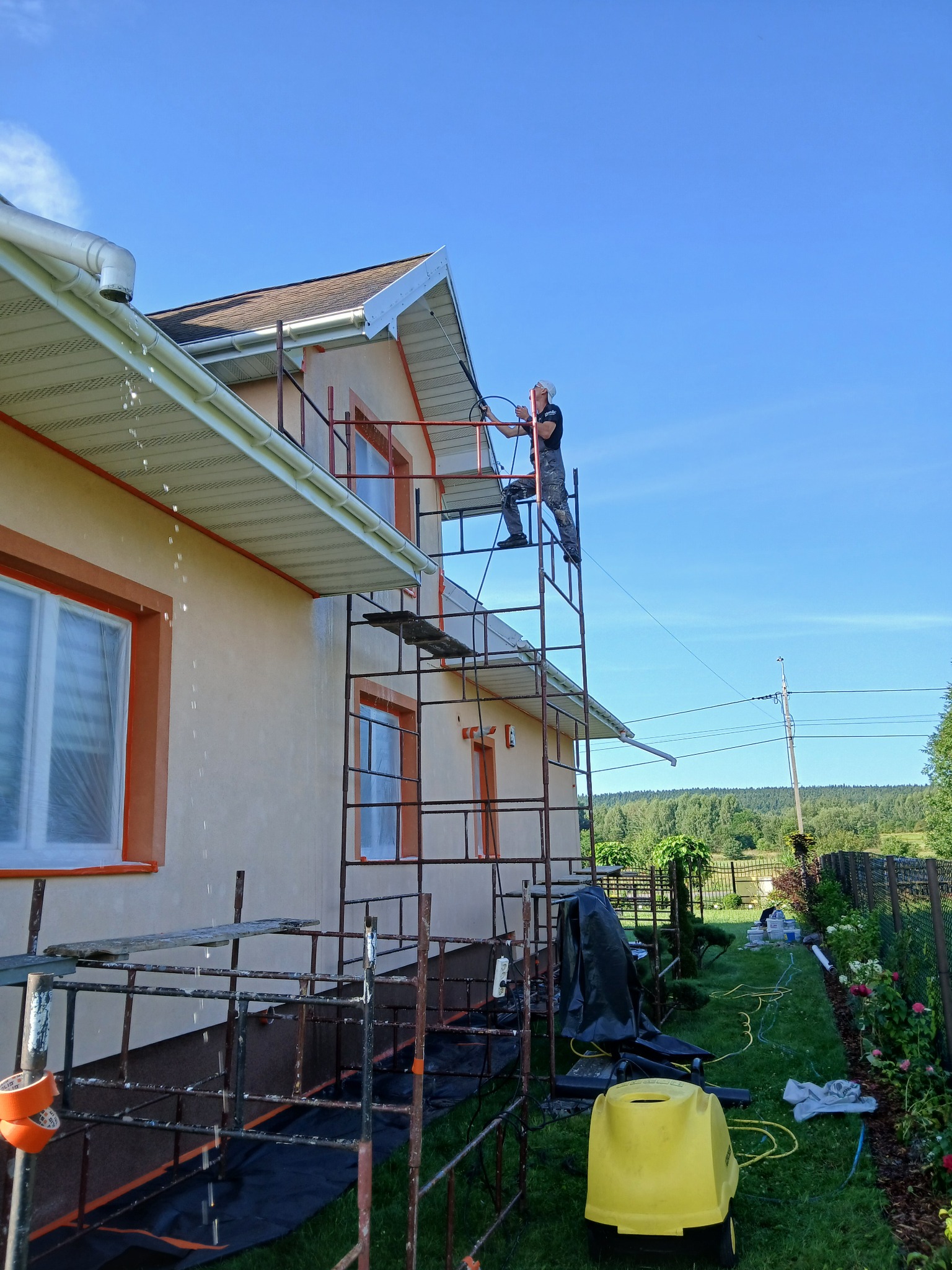 Pracownik myje elewację domu z rusztowania przy użyciu myjki ciśnieniowej. W tle zielony krajobraz i błękitne niebo. Widoczne detale elewacji i narzędzia pracy.