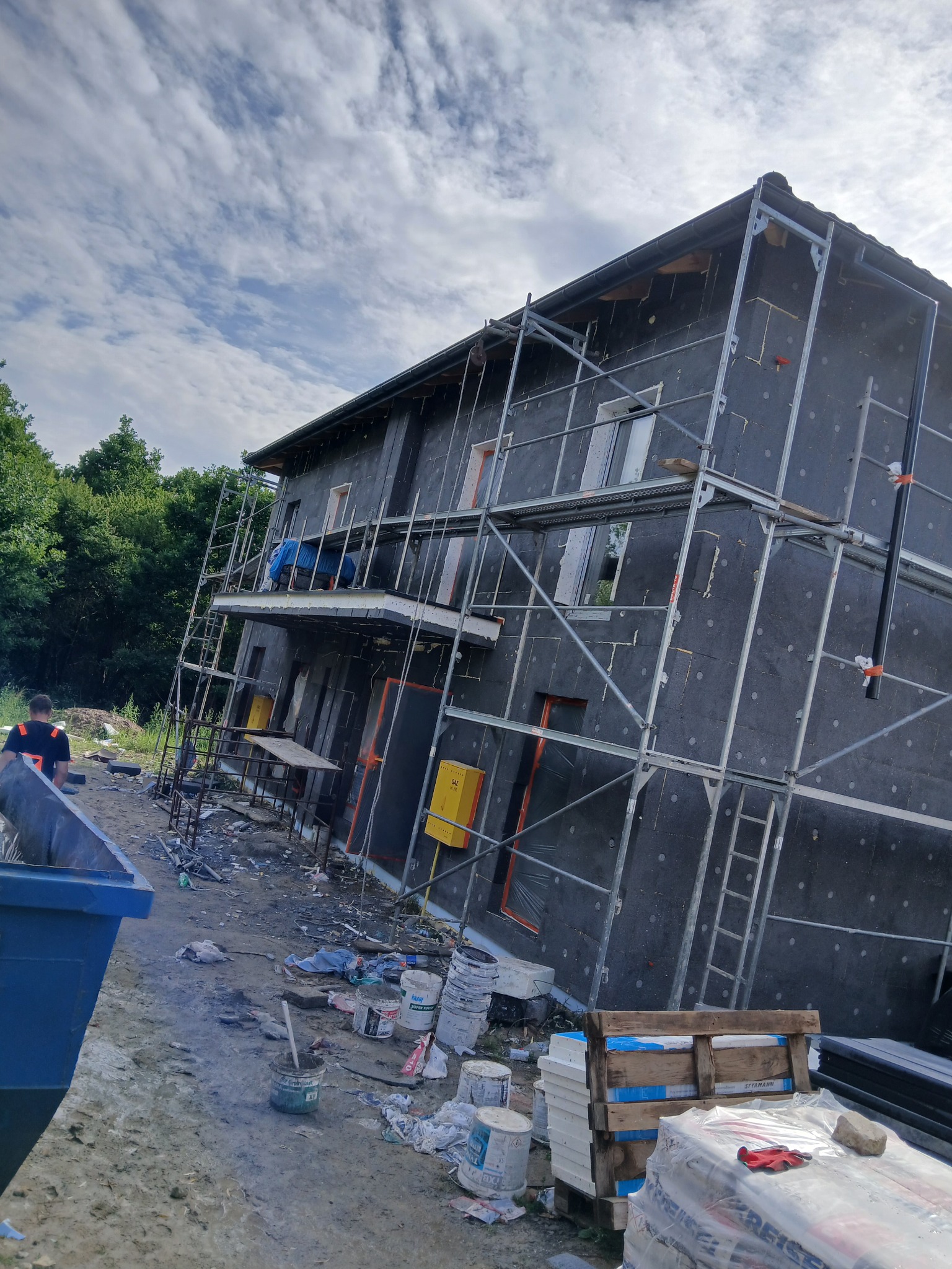 Dom w trakcie ocieplania elewacji, widoczne rusztowania, materiały izolacyjne i pracownik w tle. Etap prac przed nałożeniem tynku.