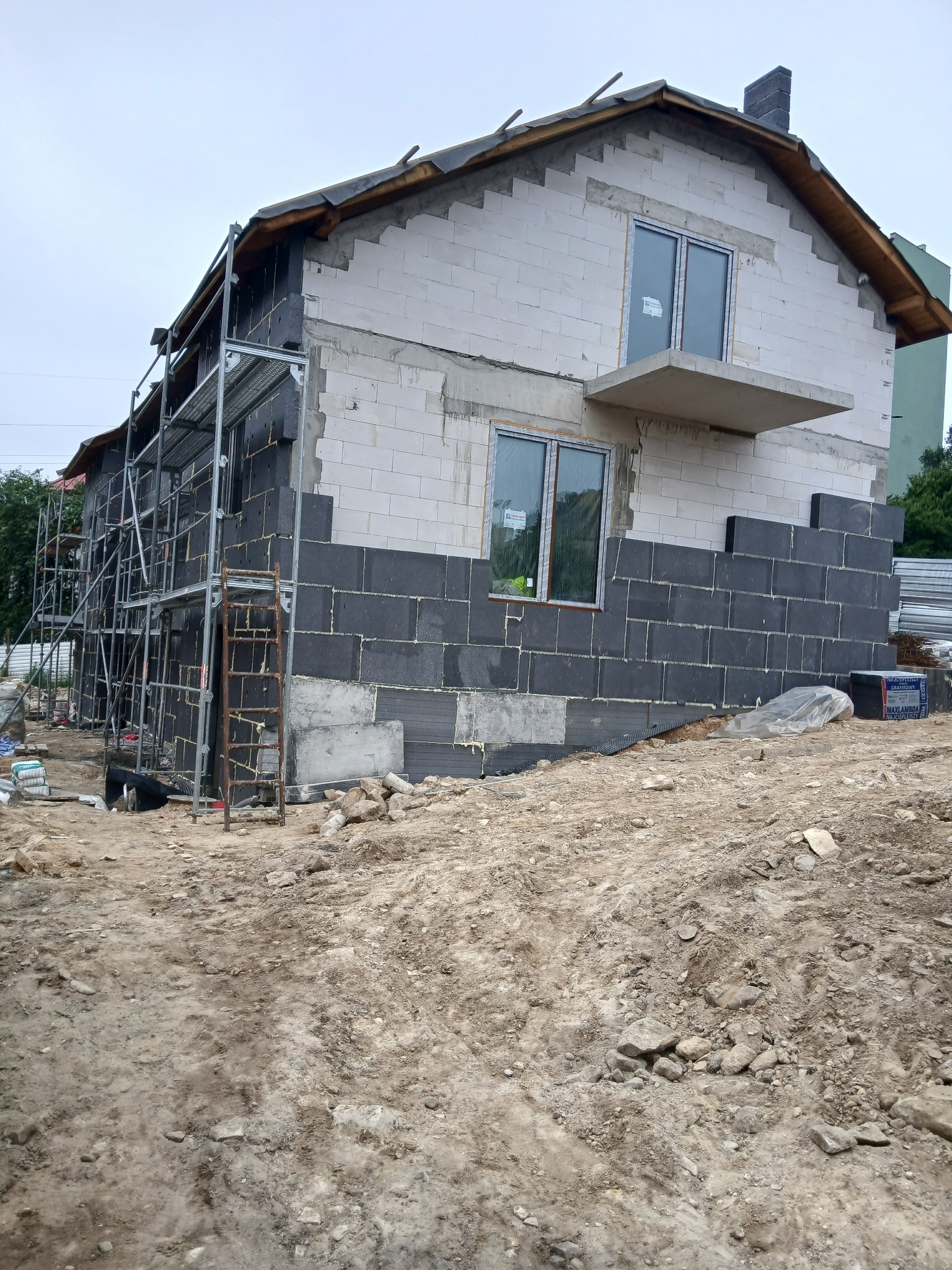 Budowa domu w toku: częściowo ocieplona elewacja grafitowym styropianem, rusztowania przy ścianie, widoczne bloczki z betonu komórkowego na górze.
