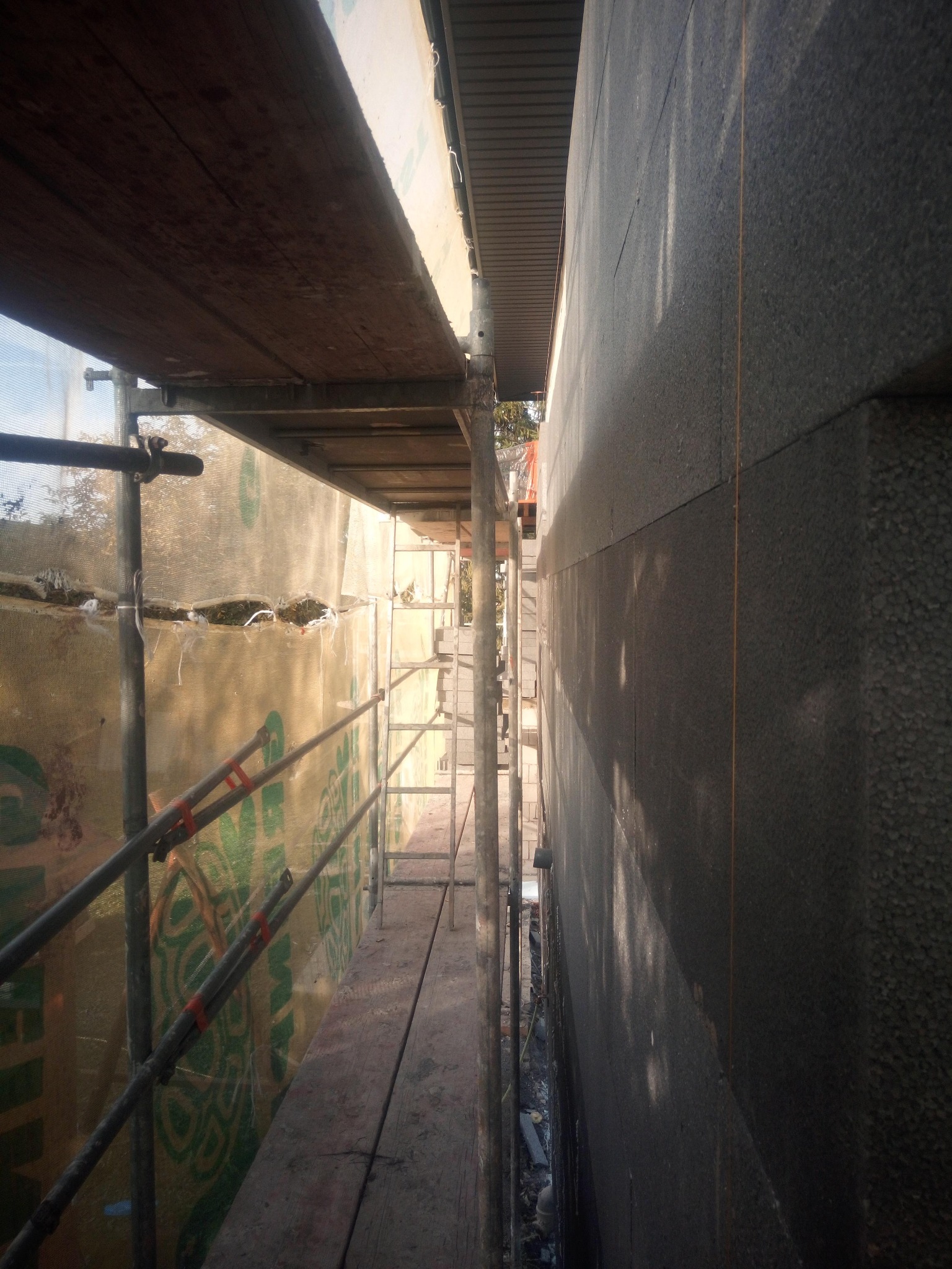 Elewacja budynku w trakcie ocieplania, widok z rusztowania. Szare płyty izolacyjne, siatka ochronna z zielonym wzorem. Wąski kadr z bliska.