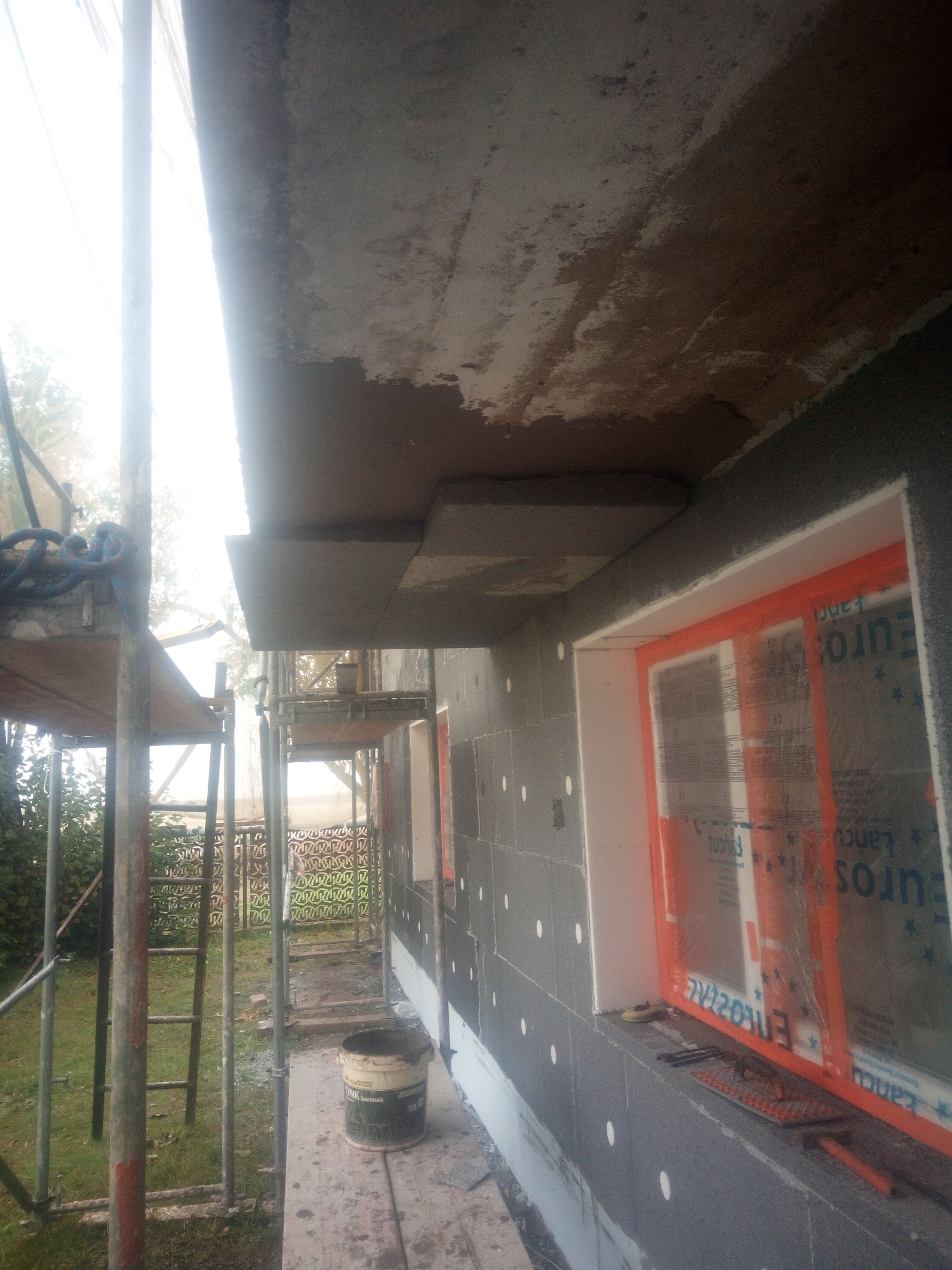 Elewacja budynku w trakcie ocieplania, widoczne rusztowania, styropian grafitowy z kołkami, okno zabezpieczone folią. Ujęcie z dołu, w tle zieleń.