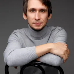 Portret mężczyzny w szarym golfie, siedzącego na czarnym krześle na tle szarego studia. Uśmiech, delikatny zarost, ręce oparte o oparcie krzesła.