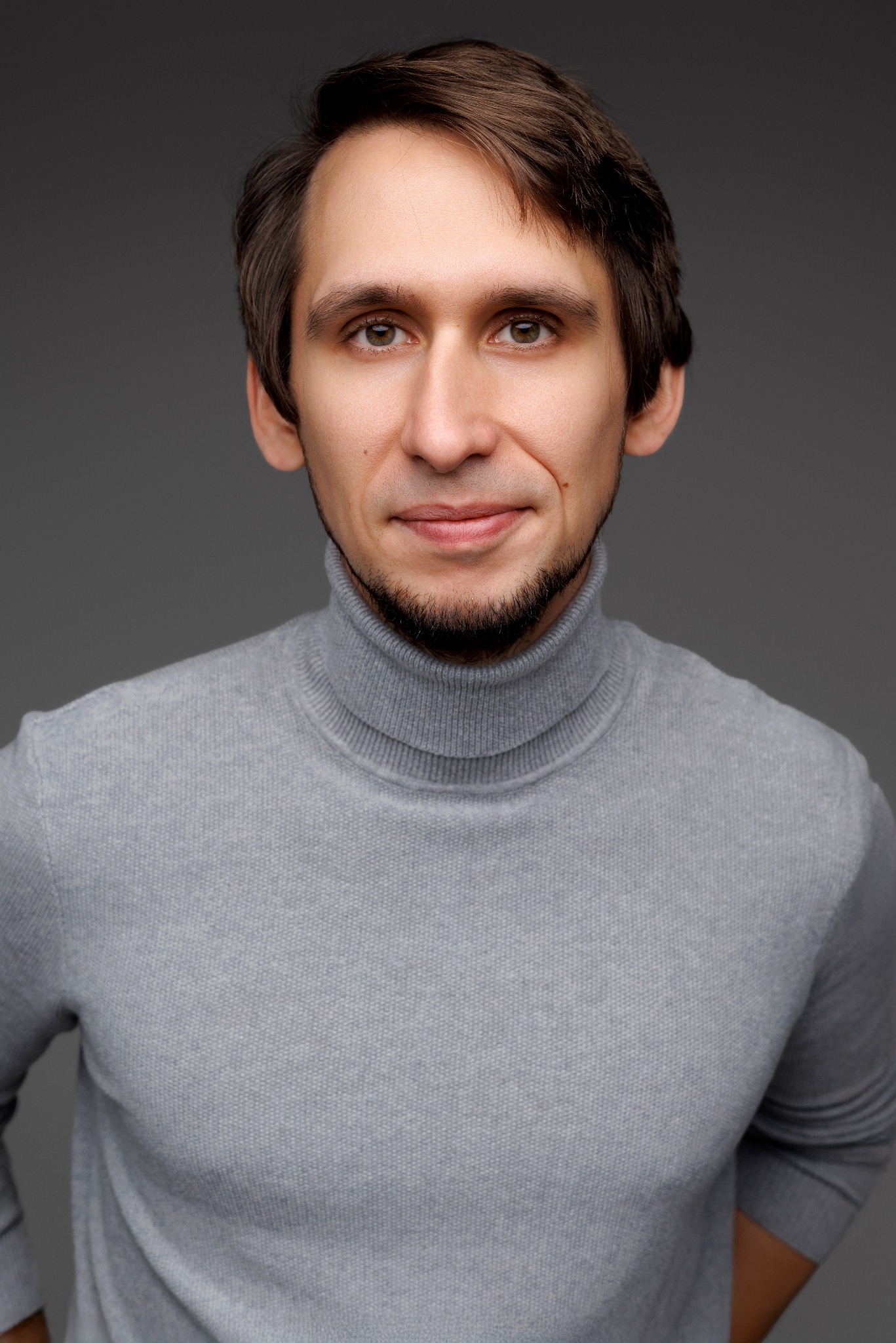 Portret mężczyzny w szarym golfie na tle szarego tła. Uśmiechnięty mężczyzna z brązowymi włosami i zarostem patrzy wprost w obiektyw. Naturalne oświetlenie.