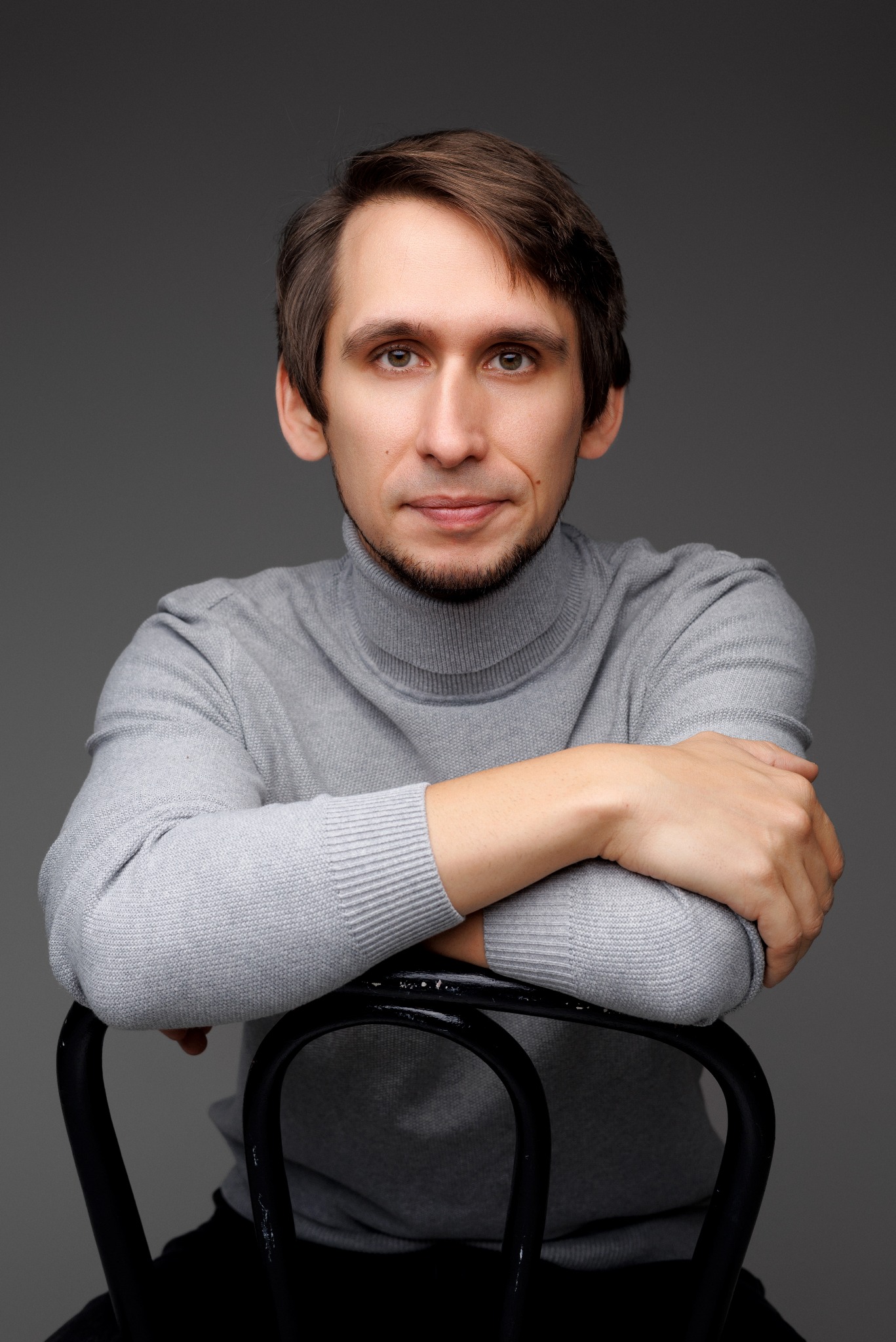 Portret mężczyzny w szarym golfie, siedzącego na czarnym krześle na tle szarego studia. Uśmiech, delikatny zarost, ręce oparte o oparcie krzesła.