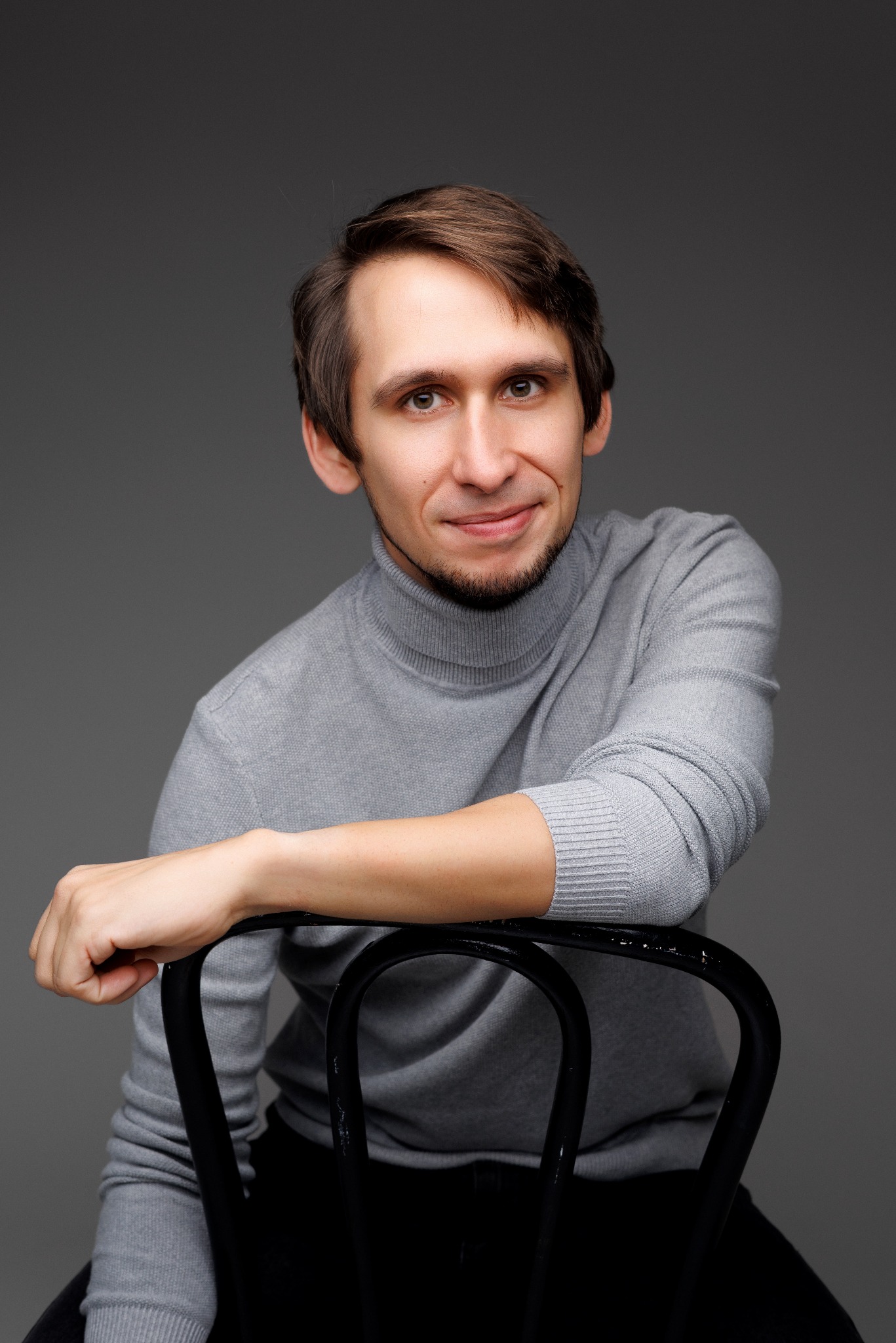 Portret uśmiechniętego mężczyzny w szarym golfie, opierającego się o czarne krzesło na jednolitym szarym tle. Ujęcie od przodu, z lekkim uśmiechem i widocznym zarostem.