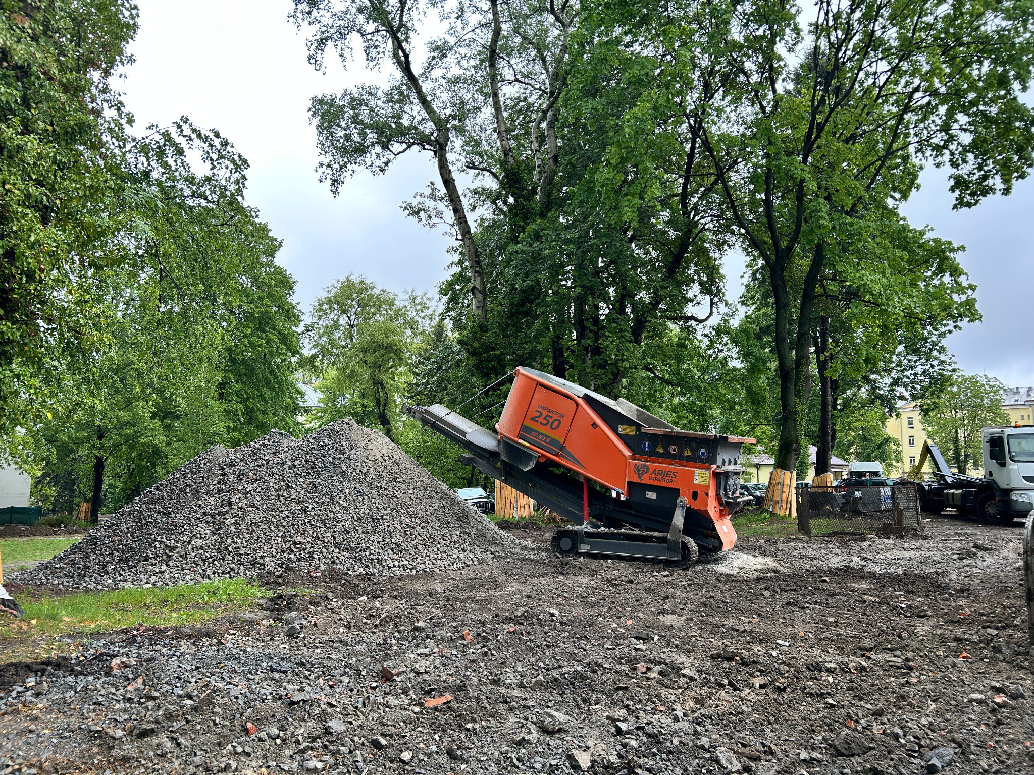 Pomarańczowa kruszarka Aries Impaktori 250 RUB II obok sterty gruzu na placu budowy, otoczona zielenią drzew. W tle widoczny budynek i ciężarówka.
