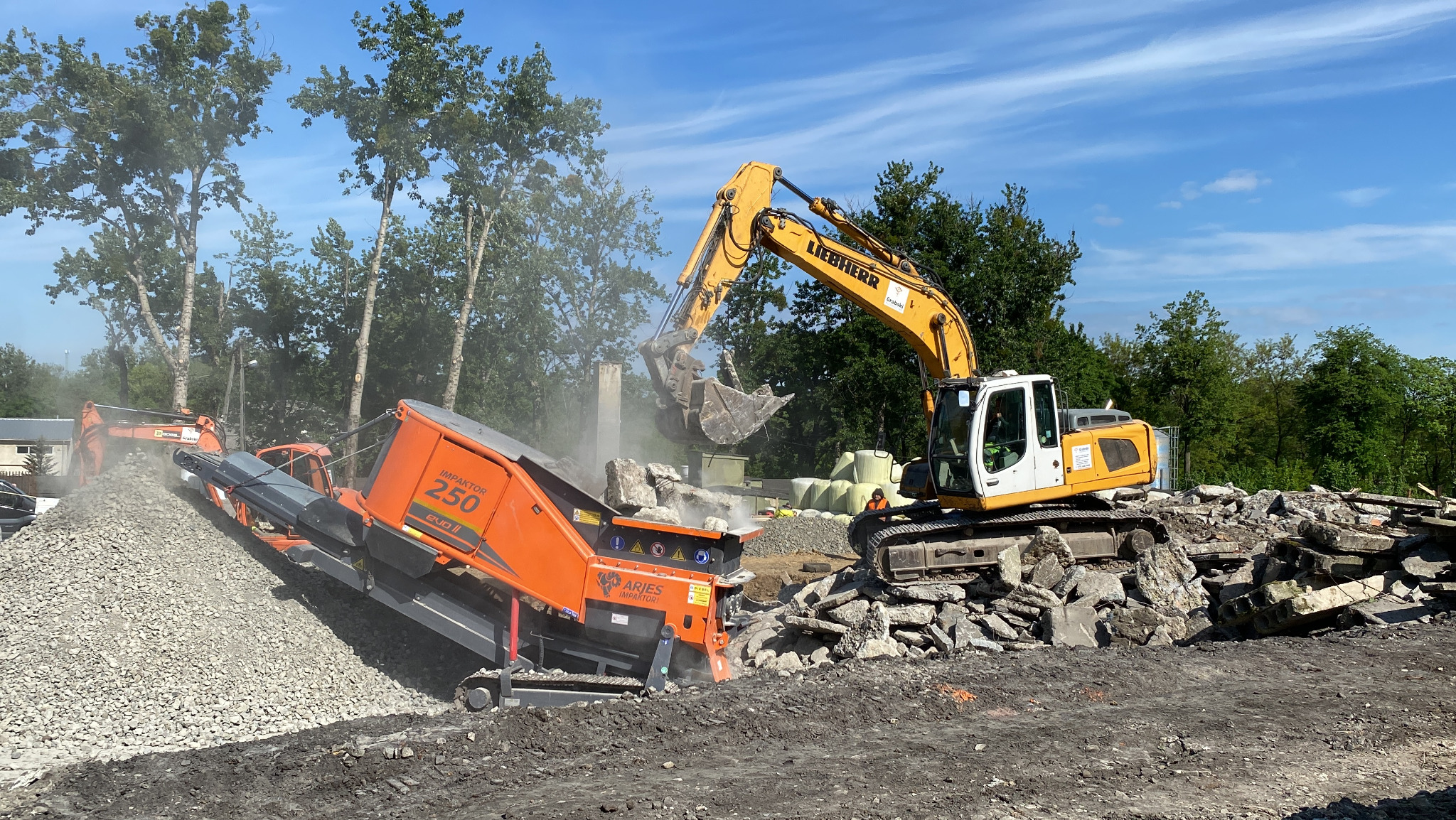 Żółta koparka Liebherr i pomarańczowa kruszarka Impaktor 250 na placu rozbiórki, z gruzem i pyłem w tle, w słoneczny dzień.