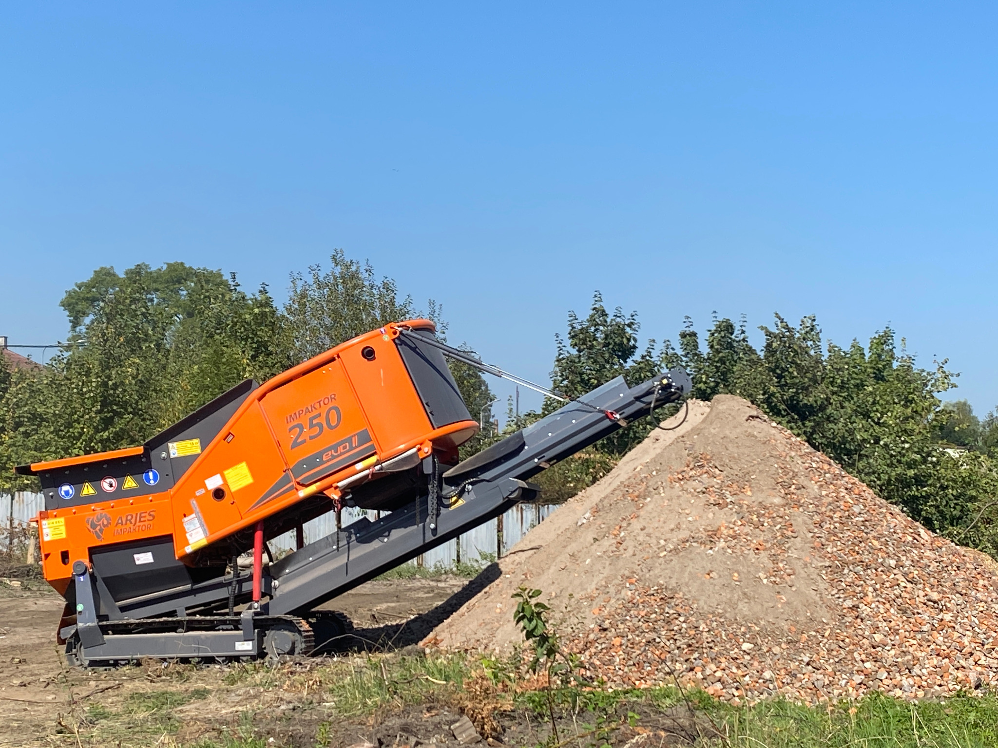 Pomarańczowa maszyna do kruszenia gruzu Aries Impaktor 250 Evo II na gąsienicach, obok hałda pokruszonego materiału. Błękitne niebo w tle.