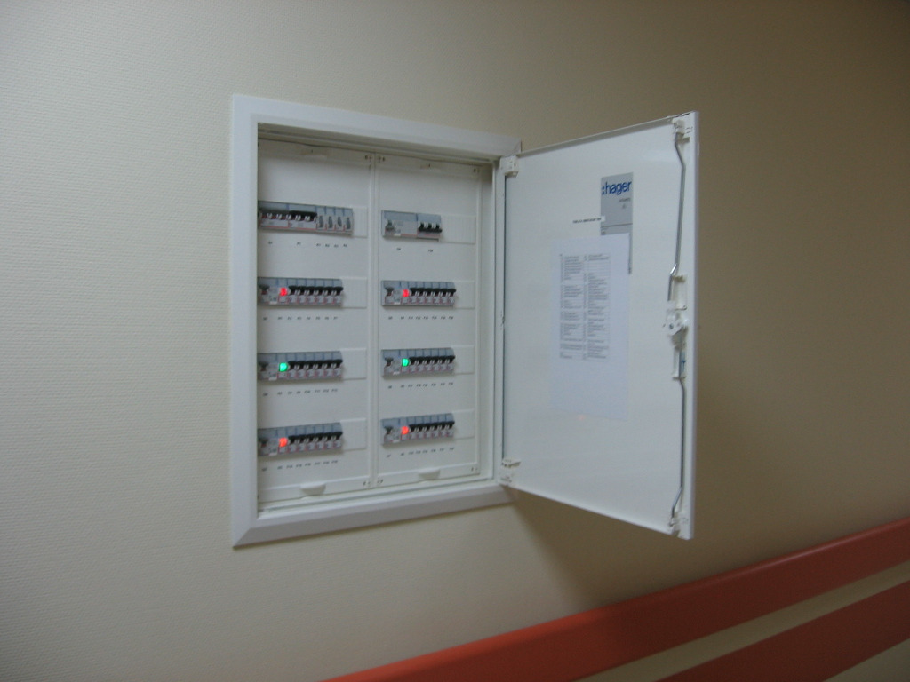 Otwarta skrzynka elektryczna Hager w ścianie, widoczne bezpieczniki z diodami. Biała obudowa, kremowe ściany i pomarańczowa barierka w tle.
