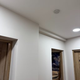 Rivano - Świeżo pomalowane, białe ściany korytarza z drewnianymi ościeżnicami drzwiowymi. Widoczny czujnik dymu na suficie i fragment tapety w pokoju za otwartymi drzwiami.