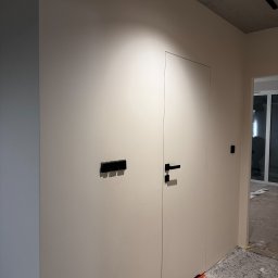 Rivano - Minimalistyczne wnętrze: gładka, beżowa ściana z ukrytymi drzwiami i czarnymi akcentami w postaci włączników i klamki. Widoczne detale wykończeniowe.