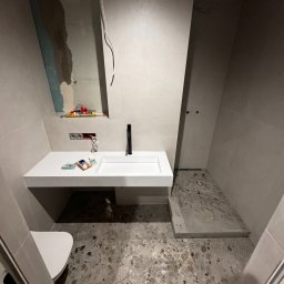 Rivano - Nowoczesna łazienka w trakcie wykańczania: biała umywalka z czarną armaturą, płytki lastryko na podłodze, widoczne narzędzia i instalacje elektryczne.