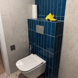 Rivano - Nowoczesna toaleta podwieszana z bidetką, obudowana ciemnoniebieskimi, pionowo ułożonymi płytkami. Na obudowie leży żółta ściereczka i rolka papieru. Ściany wykończone szarą płytką.