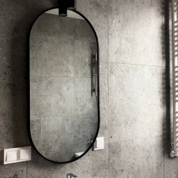 Rivano - Szara łazienka z owalnym lustrem z czarną ramą i nowoczesną armaturą. Widoczne płytki imitujące kamień i biały grzejnik drabinkowy.