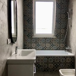 Rivano - Wykończona łazienka z wanną obudowaną dekoracyjnymi płytkami, umywalką z szafką i owalnym lustrem. Szare ściany i podłoga, okno nad wanną. Nowoczesny design.