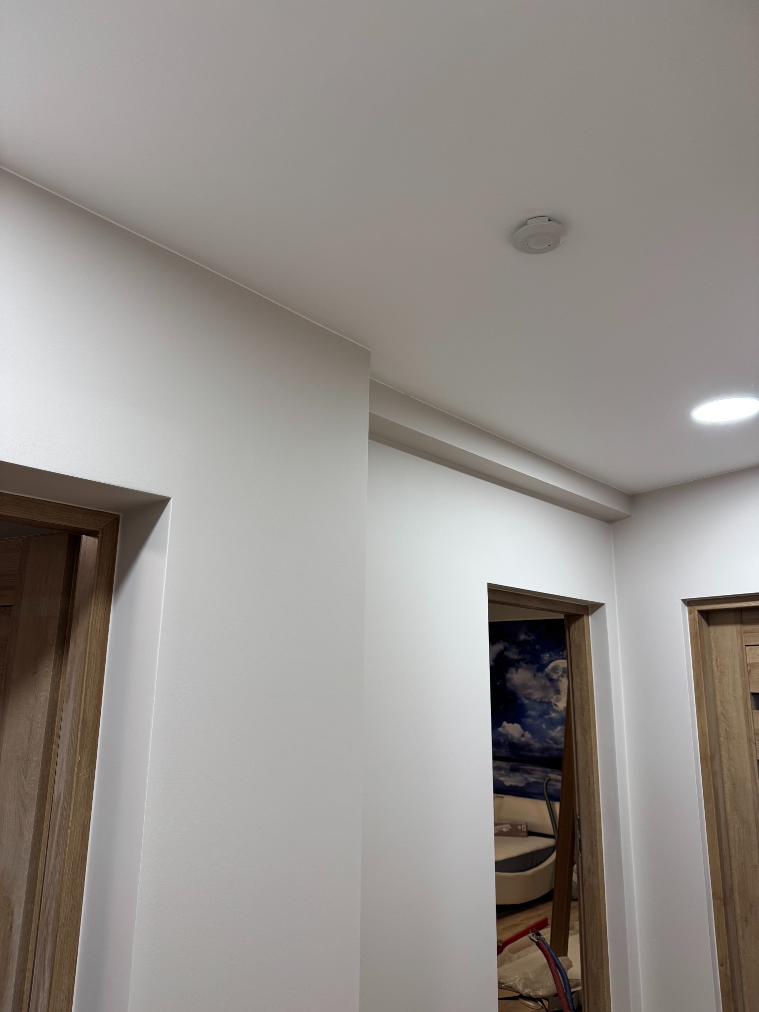 Świeżo pomalowane, białe ściany korytarza z drewnianymi ościeżnicami drzwiowymi. Widoczny czujnik dymu na suficie i fragment tapety w pokoju za otwartymi drzwiami.