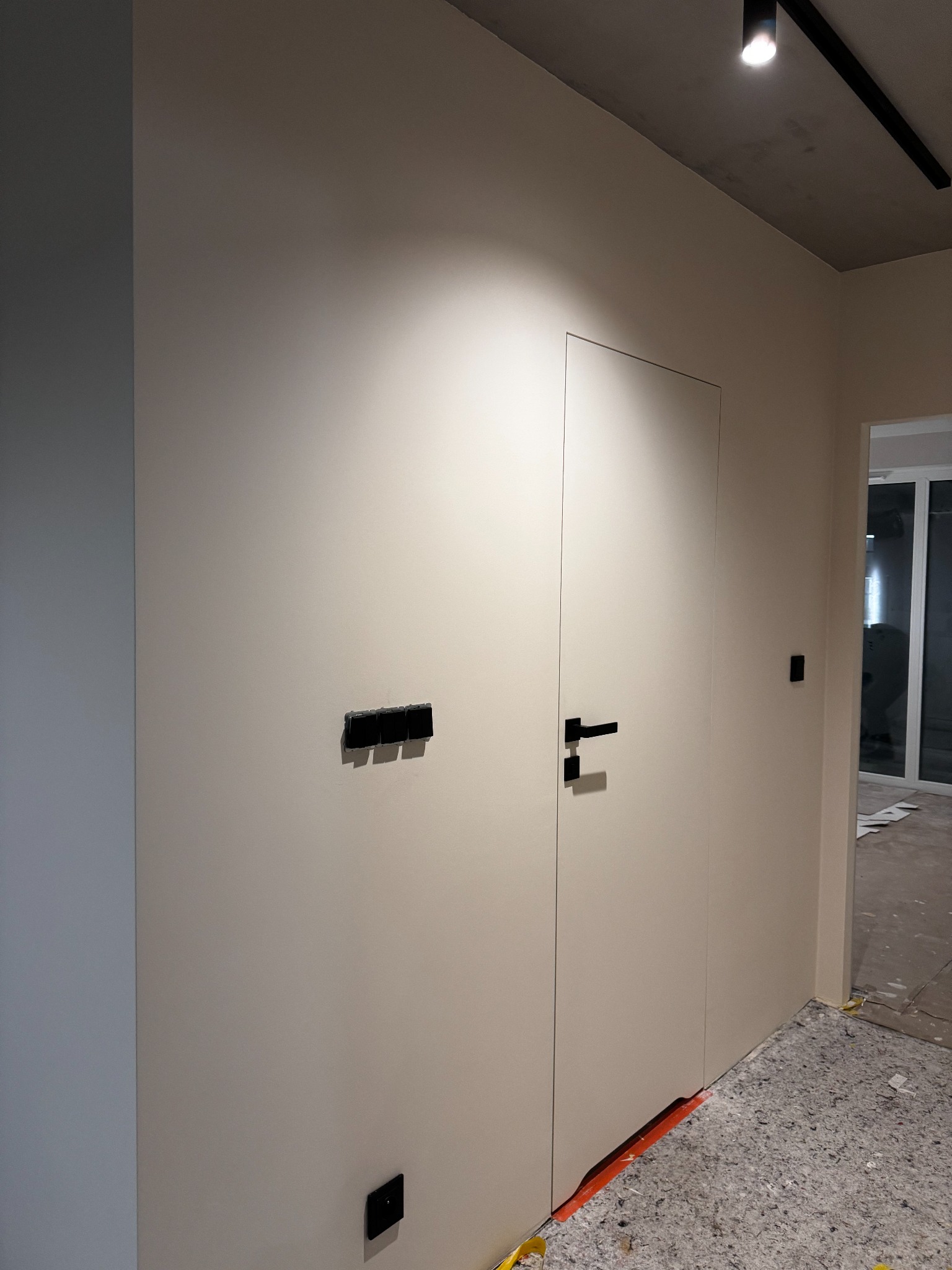 Minimalistyczne wnętrze: gładka, beżowa ściana z ukrytymi drzwiami i czarnymi akcentami w postaci włączników i klamki. Widoczne detale wykończeniowe.