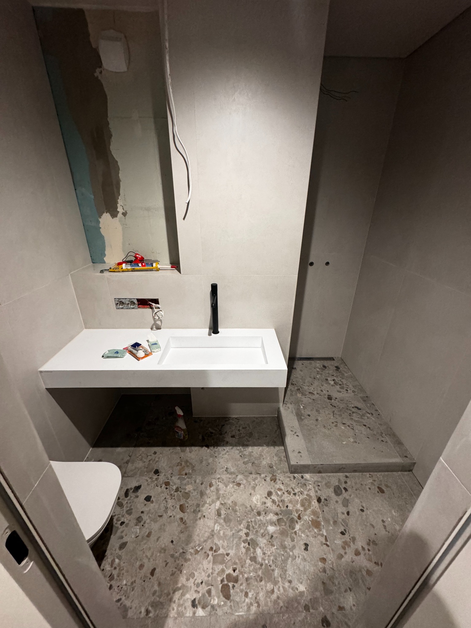 Nowoczesna łazienka w trakcie wykańczania: biała umywalka z czarną armaturą, płytki lastryko na podłodze, widoczne narzędzia i instalacje elektryczne.