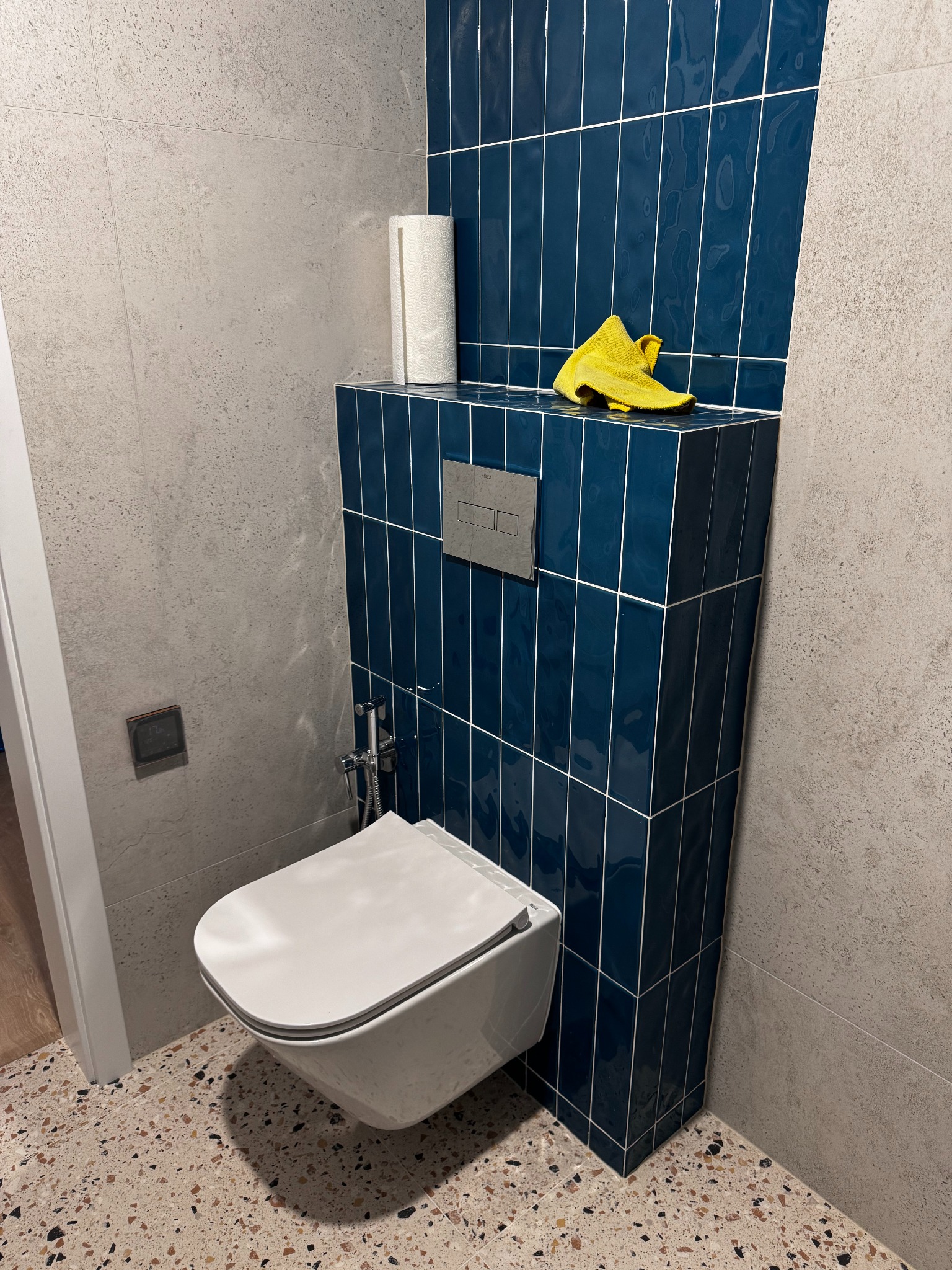 Nowoczesna toaleta podwieszana z bidetką, obudowana ciemnoniebieskimi, pionowo ułożonymi płytkami. Na obudowie leży żółta ściereczka i rolka papieru. Ściany wykończone szarą płytką.
