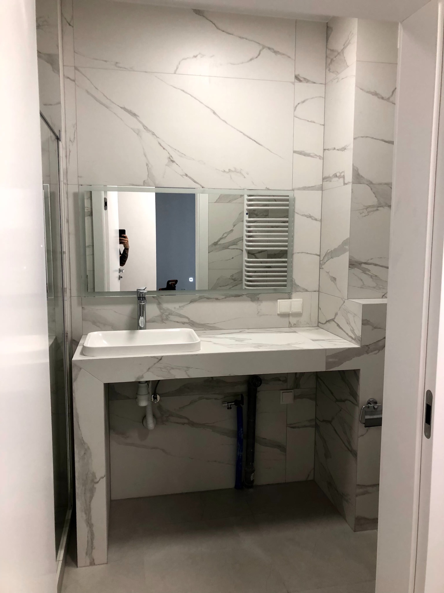 Nowoczesna łazienka z marmurowymi płytkami, prostokątną umywalką i grzejnikiem drabinkowym. W lustrze odbija się osoba robiąca zdjęcie.