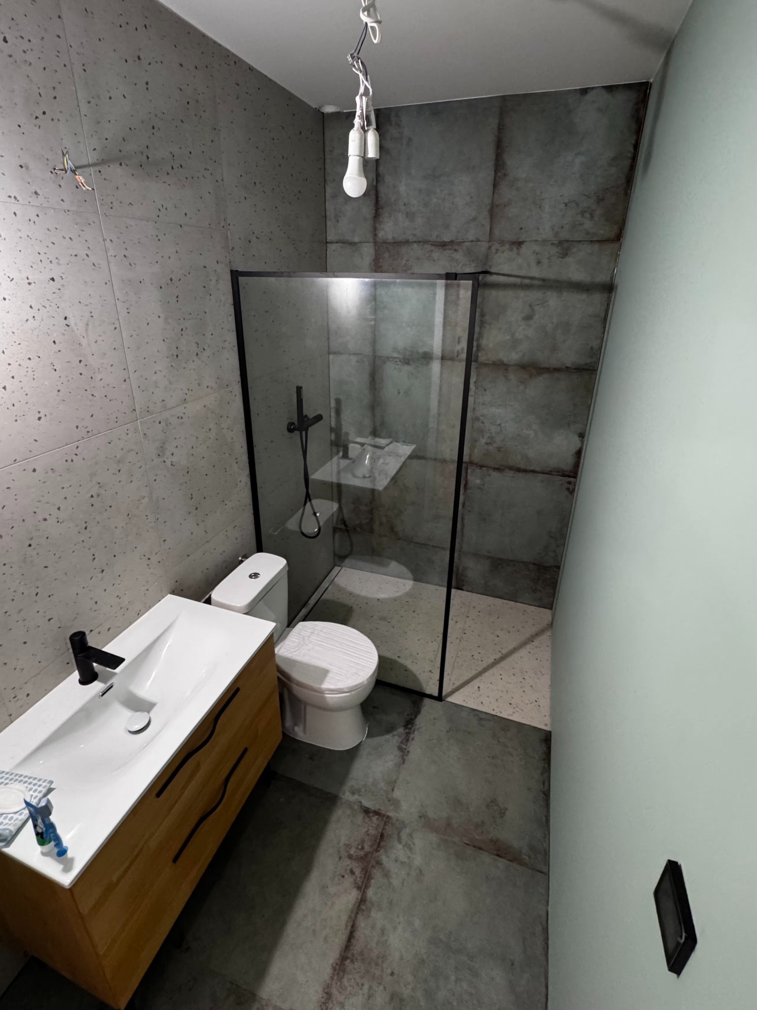 Nowoczesna łazienka z kabiną prysznicową typu walk-in, umywalką z szafką z drewnianymi frontami i toaletą. Ściany wykończone płytkami imitującymi beton.