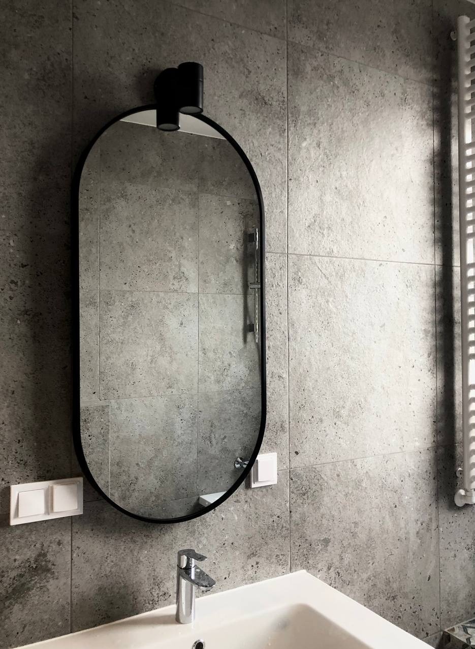 Szara łazienka z owalnym lustrem z czarną ramą i nowoczesną armaturą. Widoczne płytki imitujące kamień i biały grzejnik drabinkowy.