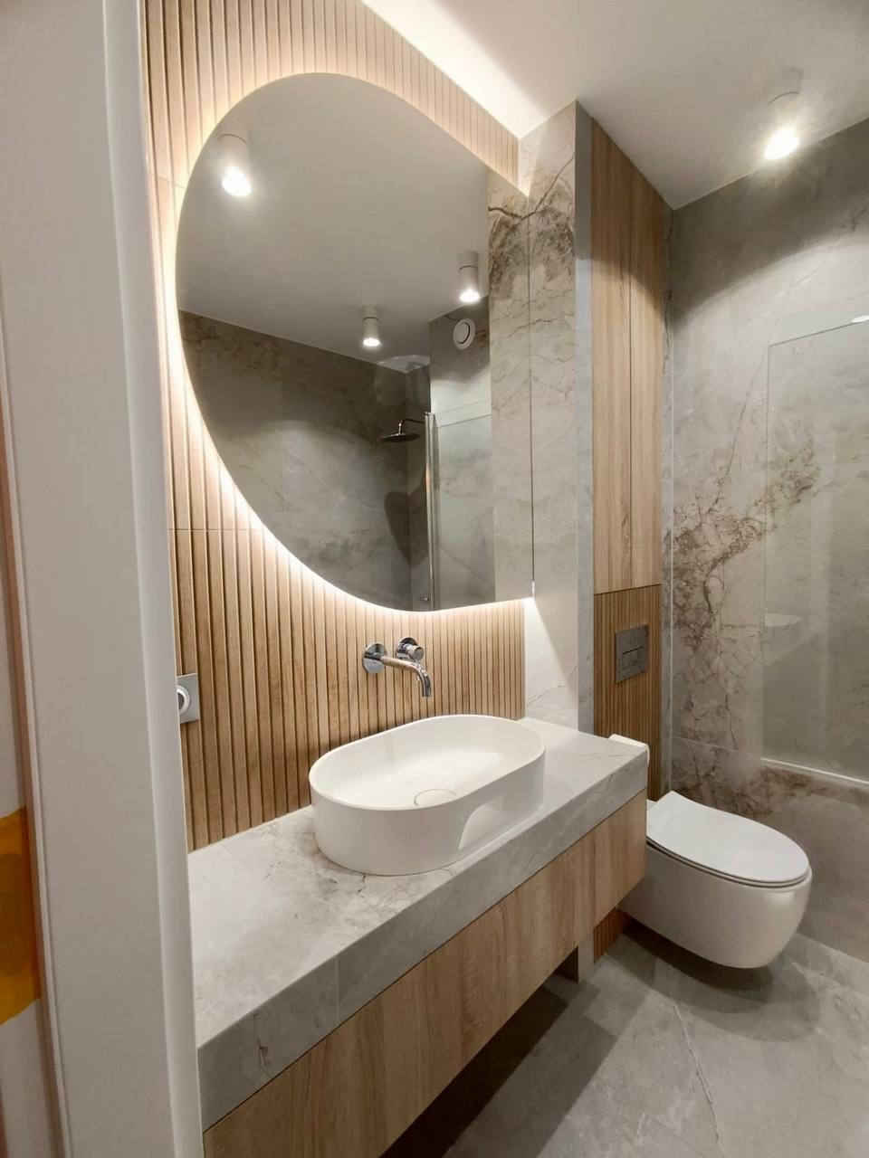 Nowoczesna łazienka z owalnym lustrem podświetlanym LED, drewnianymi panelami ściennymi i blatem z szarego marmuru. Minimalistyczna armatura i wisząca toaleta.