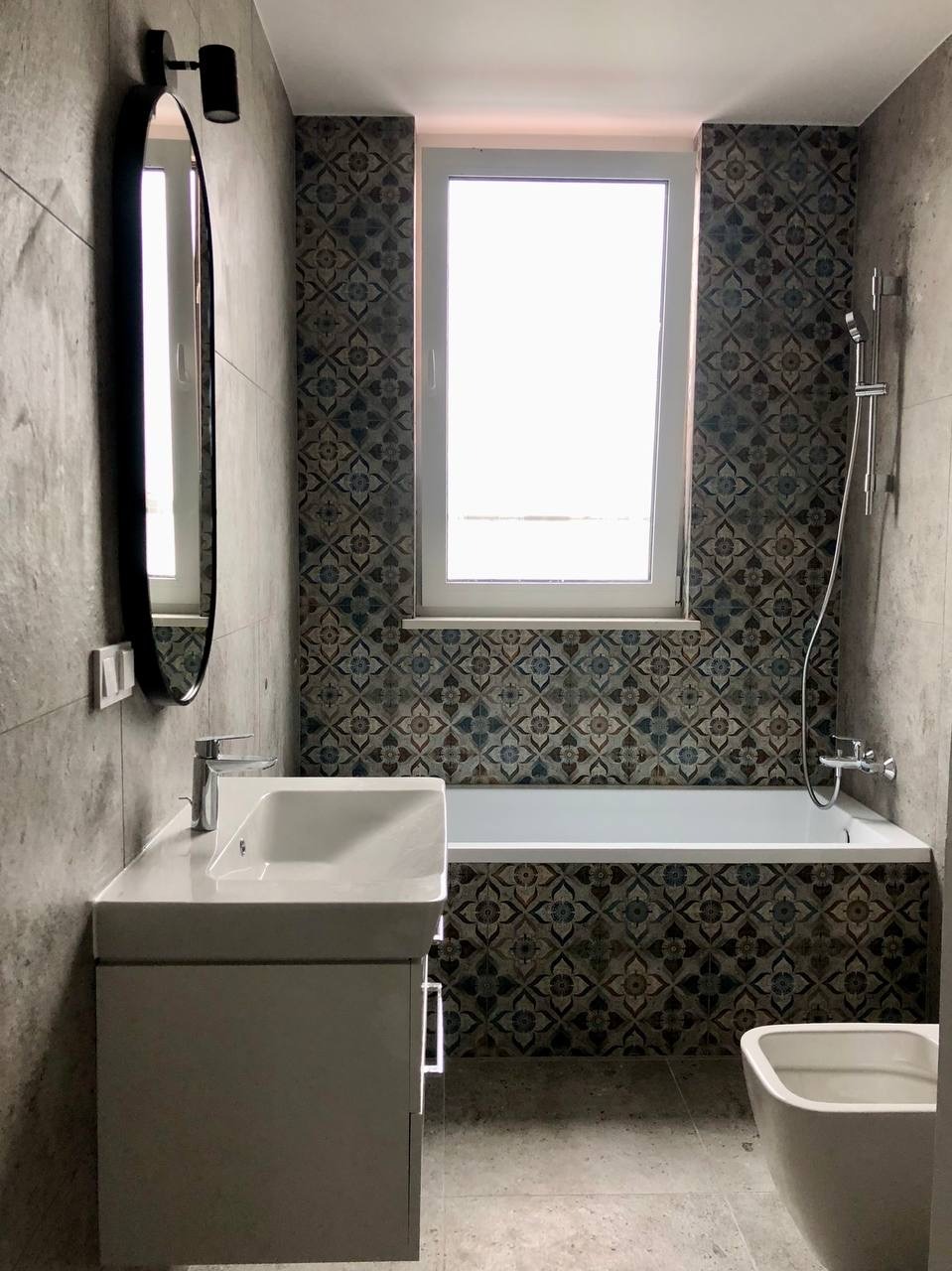 Wykończona łazienka z wanną obudowaną dekoracyjnymi płytkami, umywalką z szafką i owalnym lustrem. Szare ściany i podłoga, okno nad wanną. Nowoczesny design.