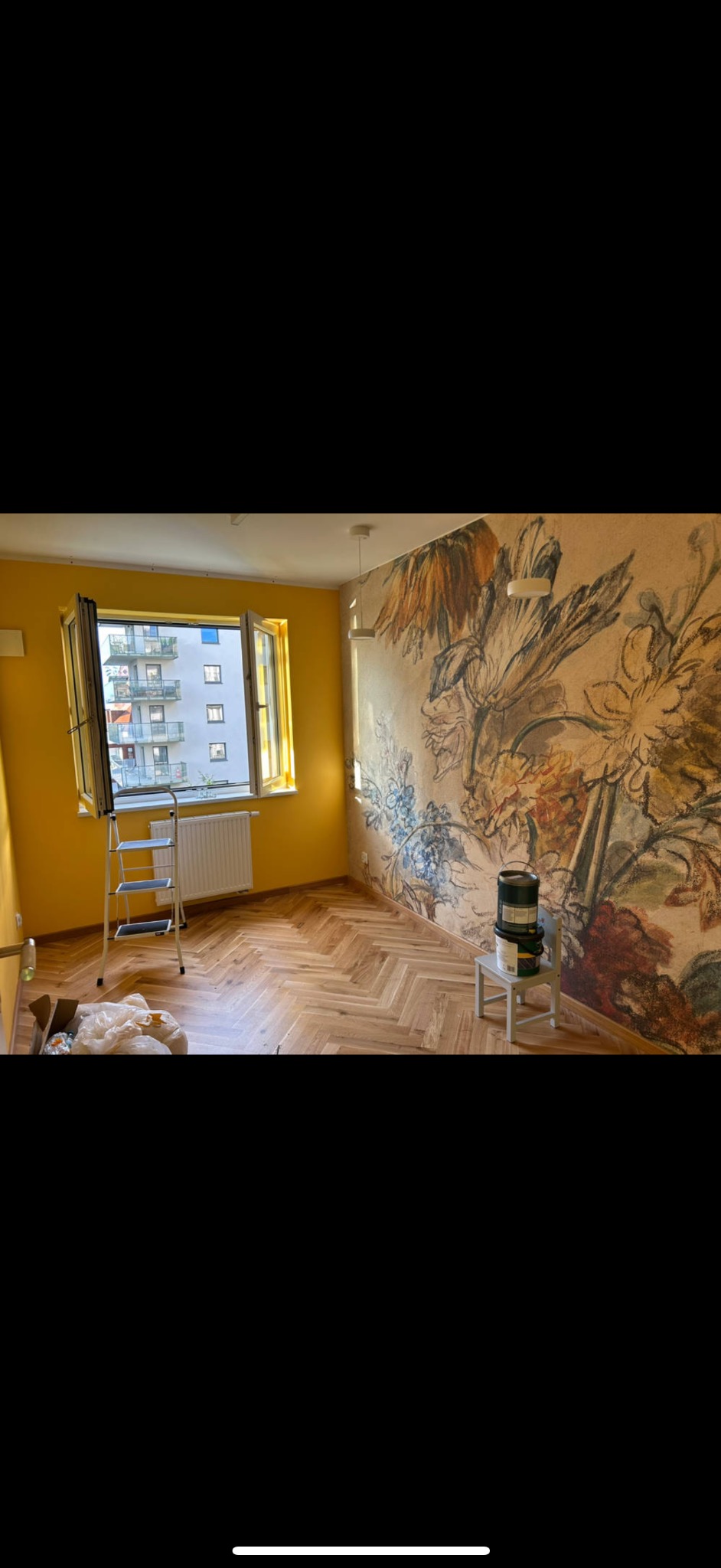 Wnętrze po malowaniu z żółtymi ścianami i tapetą w kwiaty. Widoczna drabina, podłoga w jodełkę i puszki z farbą na krześle. Nowoczesne mieszkanie po remoncie.