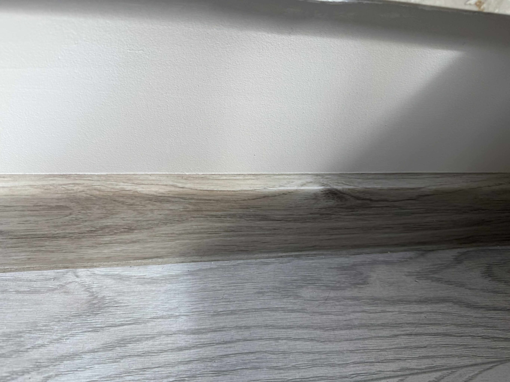 Ściana pomalowana na biało z drewnopodobną listwą przypodłogową i podłogą. Widoczne delikatne cieniowanie na ścianie. Ujęcie z dołu.