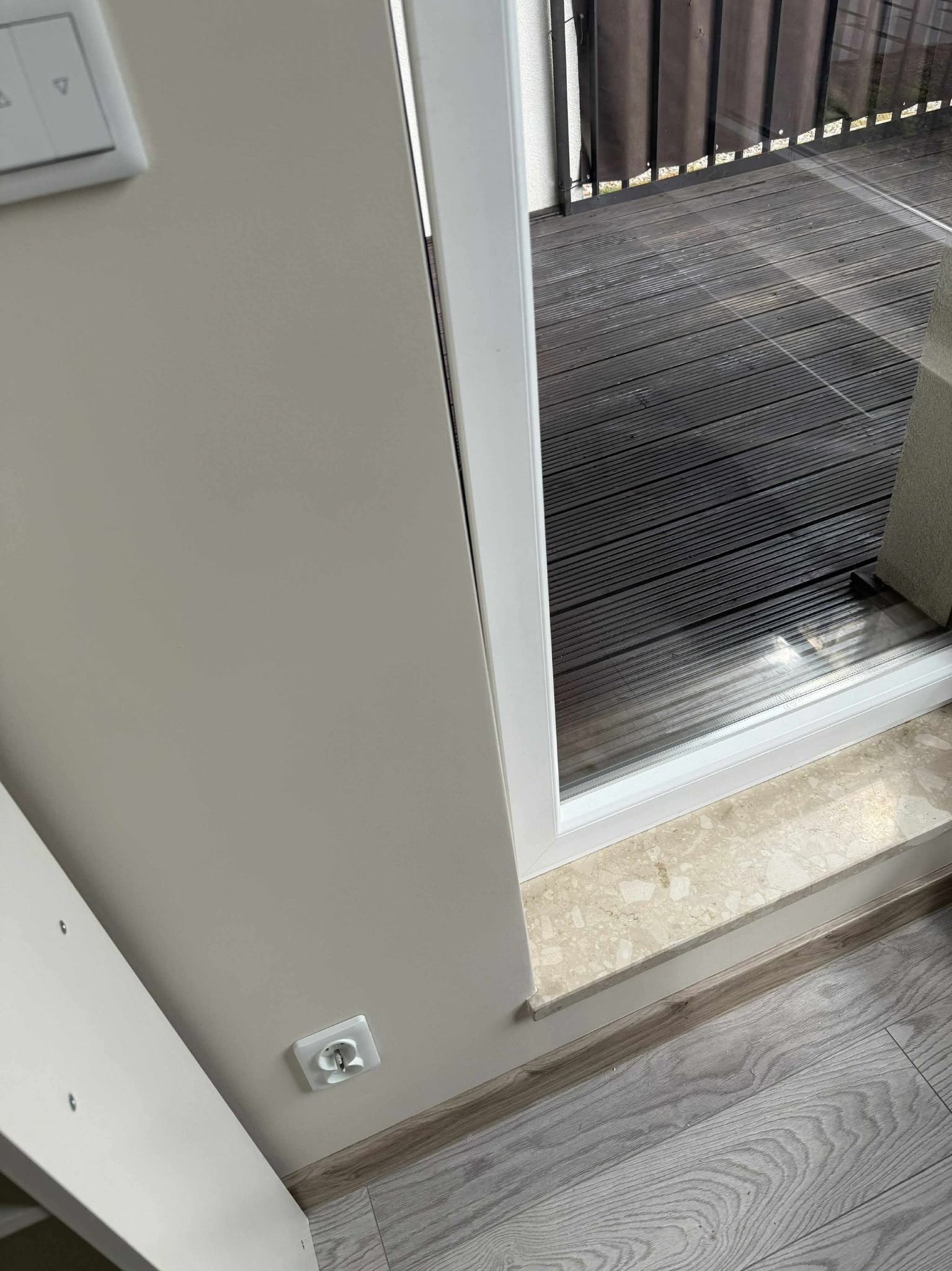 Świeżo pomalowana ściana w odcieniach beżu z widocznym gniazdkiem elektrycznym, listwą przypodłogową i oknem balkonowym z widokiem na drewniany taras.