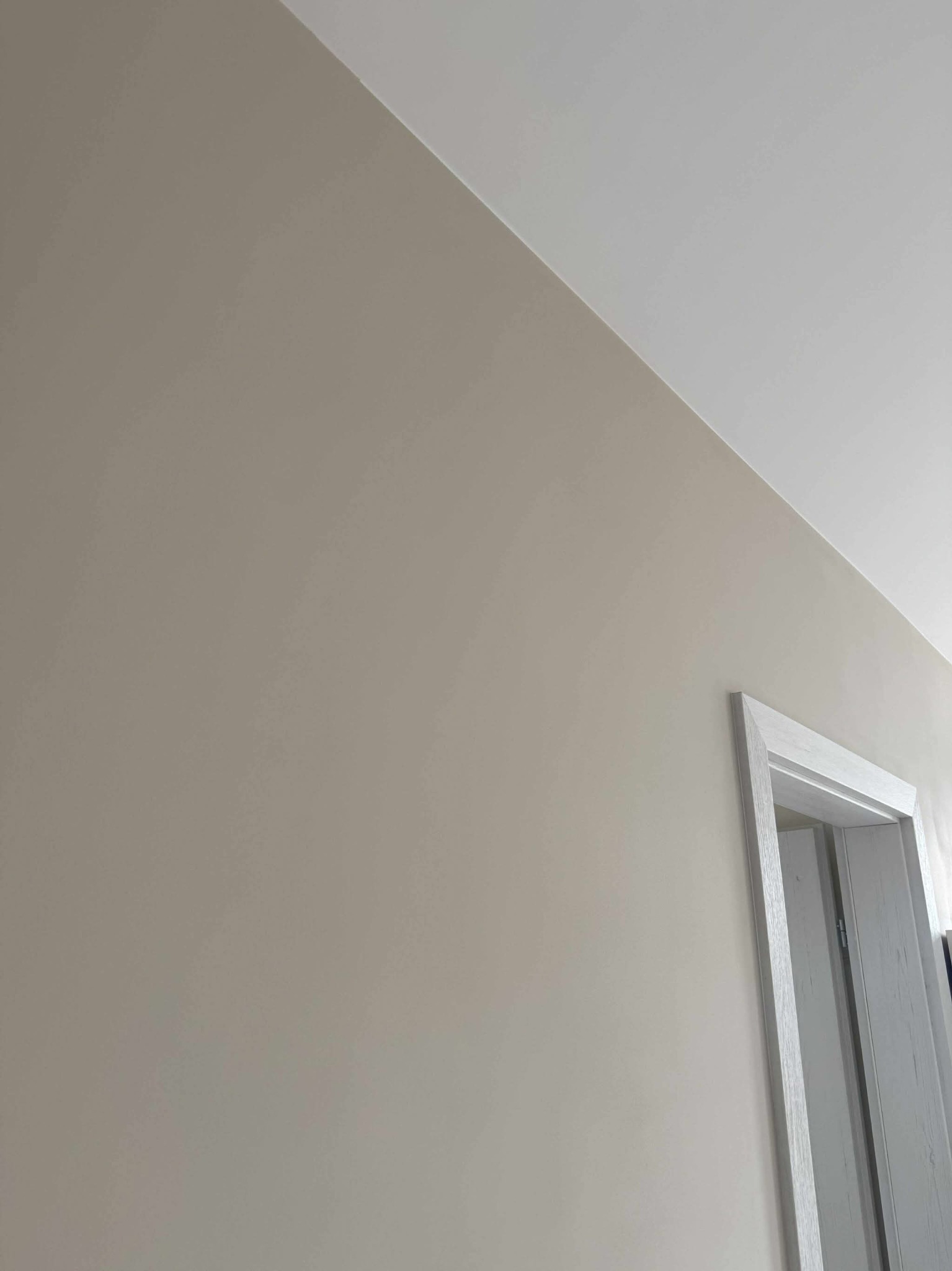 Ściana w odcieniach beżu, świeżo pomalowana, z widocznym fragmentem białej framugi drzwi. Widok z dołu, podkreślający gładką powierzchnię.