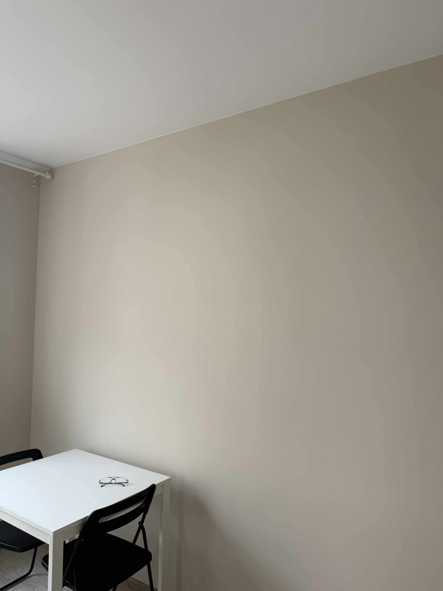 Ściana pomalowana na beżowo w pokoju z białym stołem i czarnymi krzesłami. Na stole leżą okulary.