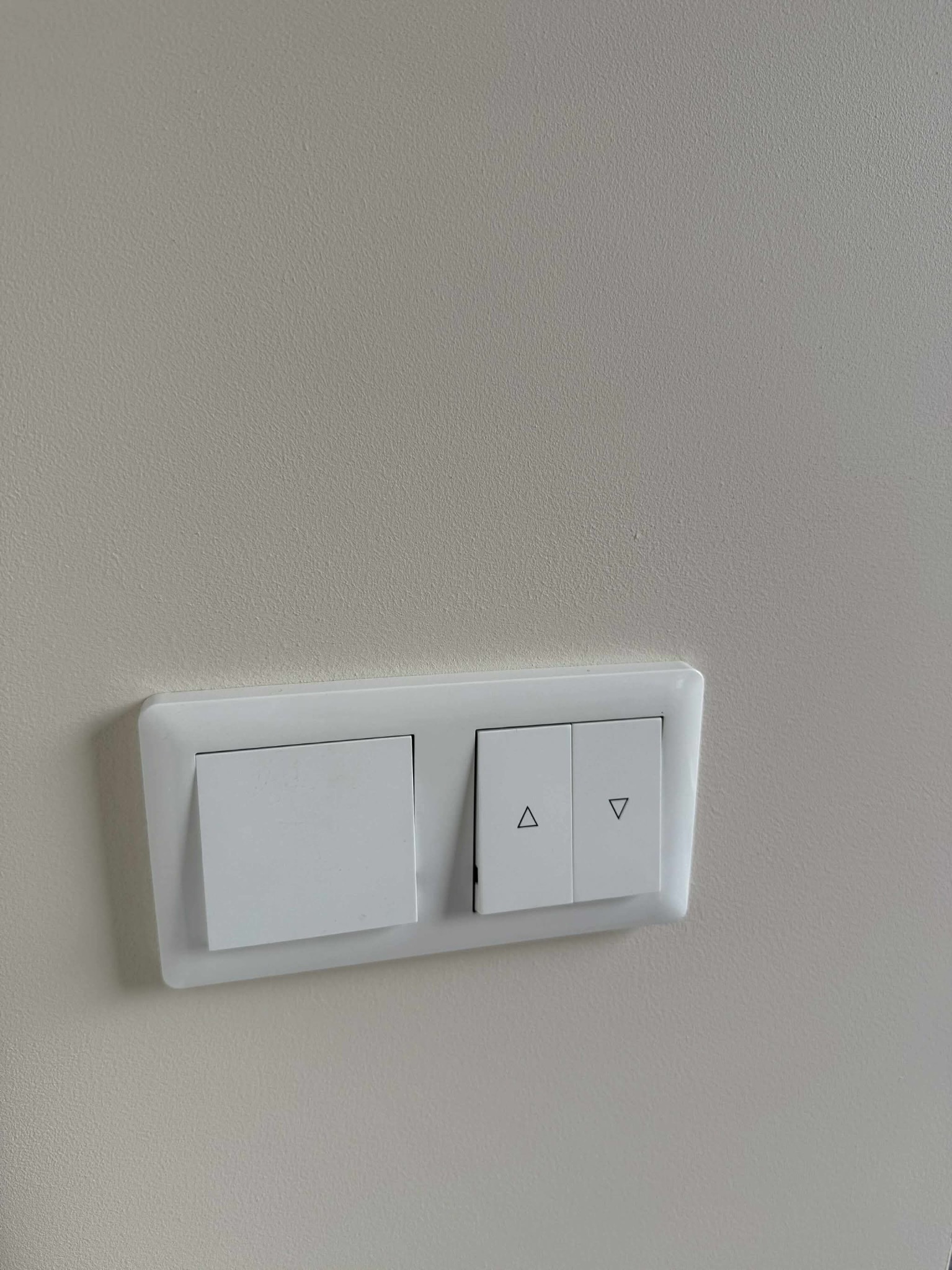 Świeżo pomalowana ściana w jasnym odcieniu z zamontowanym białym włącznikiem światła i regulatorem rolet z delikatnym cieniem.