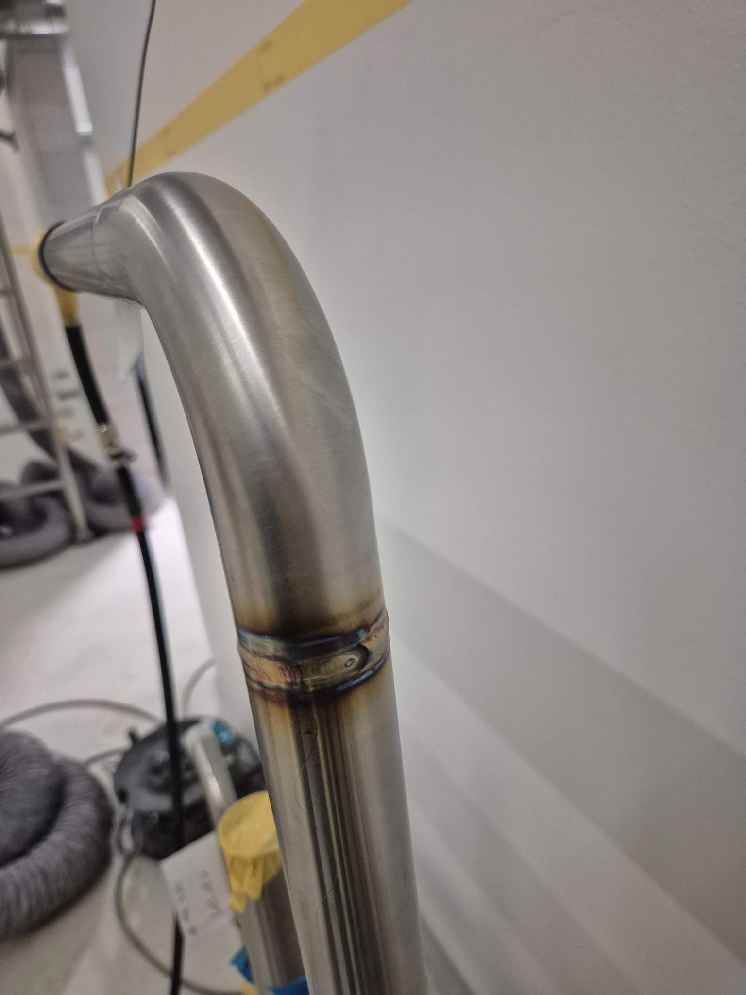 Precyzyjnie spawany, zakrzywiony element ze stali nierdzewnej, widoczne ślady spawania, surowe wnętrze warsztatu w tle, zbliżenie na łączenie.