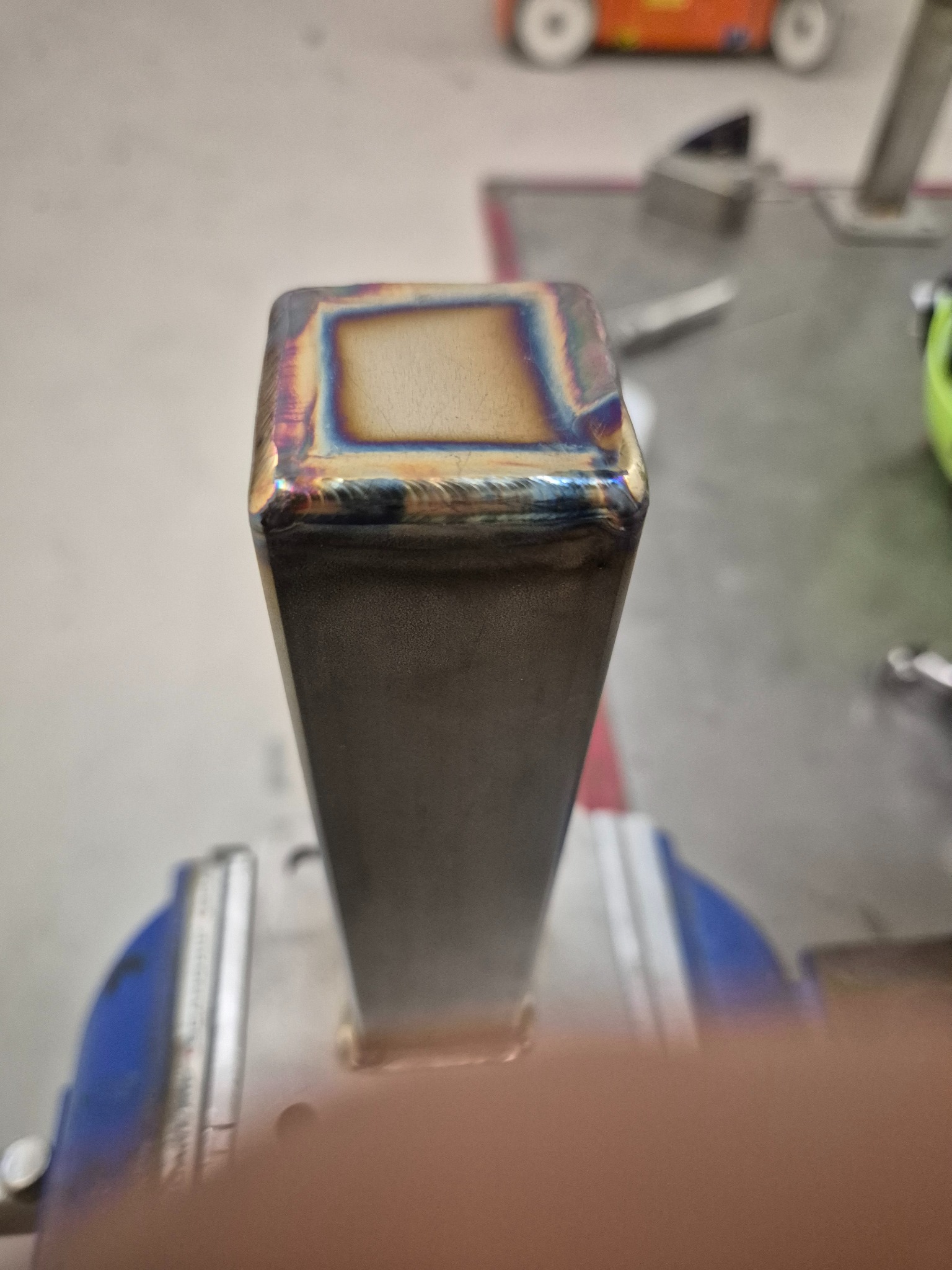 Metalowy profil kwadratowy po obróbce, widoczne ślady spawania z charakterystycznymi przebarwieniami termicznymi. Profil zamocowany w imadle.