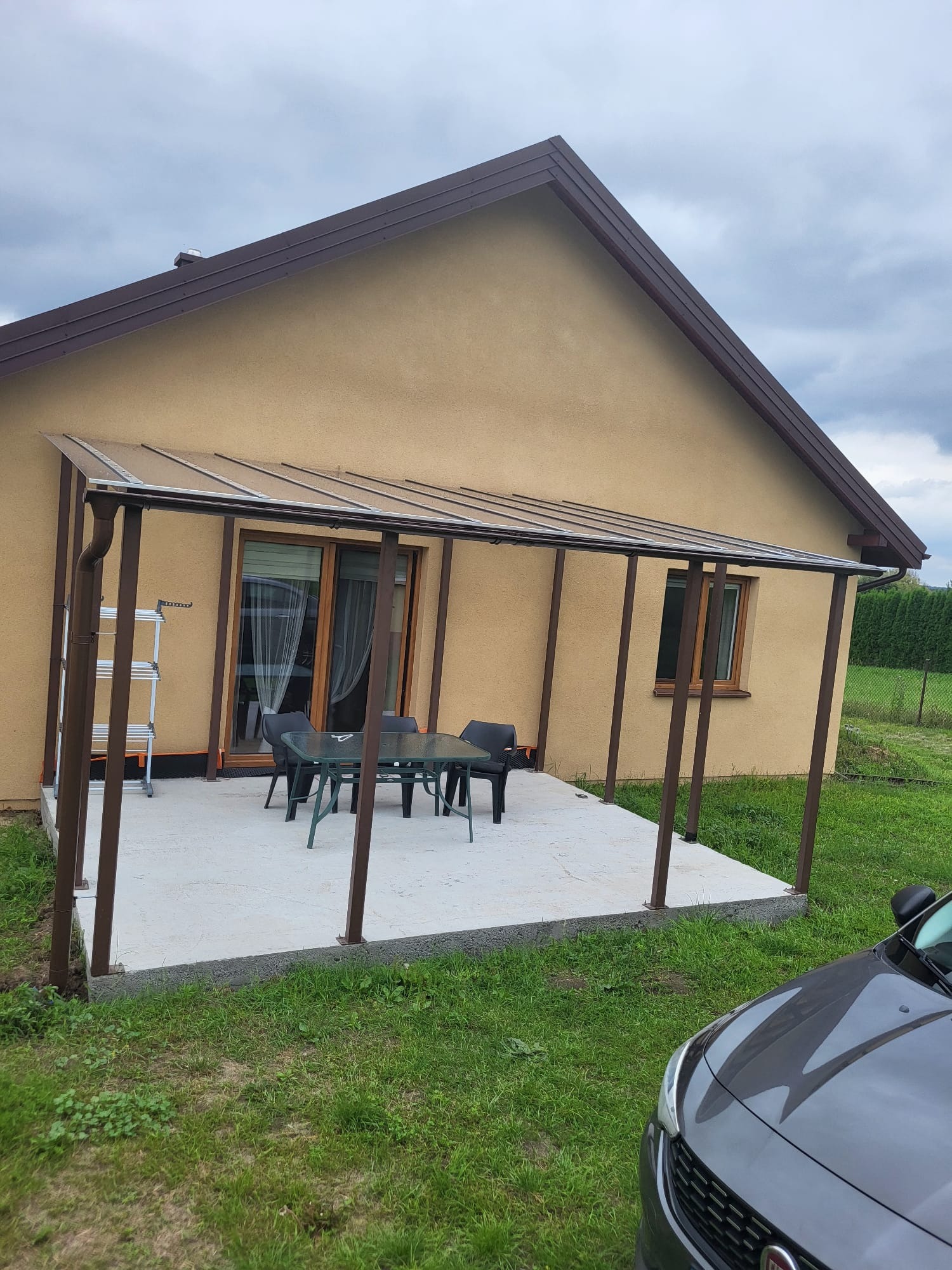 Brązowa konstrukcja z zadaszeniem przy domu, tworząca zadaszone patio z meblami ogrodowymi. Widoczny fragment samochodu i drabina oparta o ścianę.