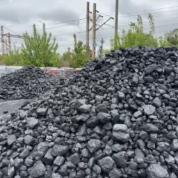 Góra czarnego węgla kamiennego na placu składowym, z widocznymi w tle workami i słupami trakcji kolejowej. Pochmurne niebo.