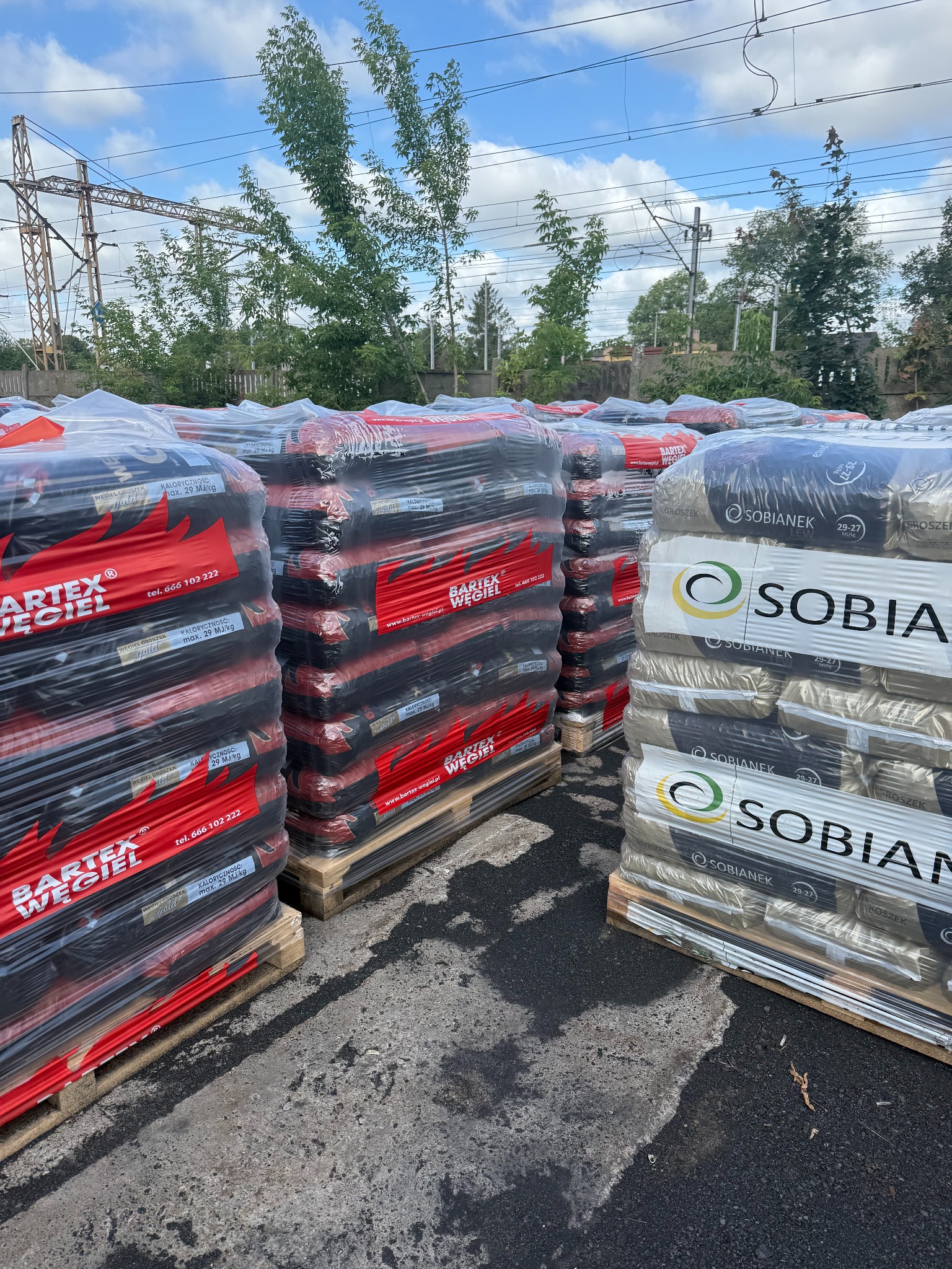 Sztaple worków z ekogroszkiem Bartex Węgiel i Sobianek na paletach, składowane na zewnątrz pod błękitnym niebem i liniami wysokiego napięcia w tle.